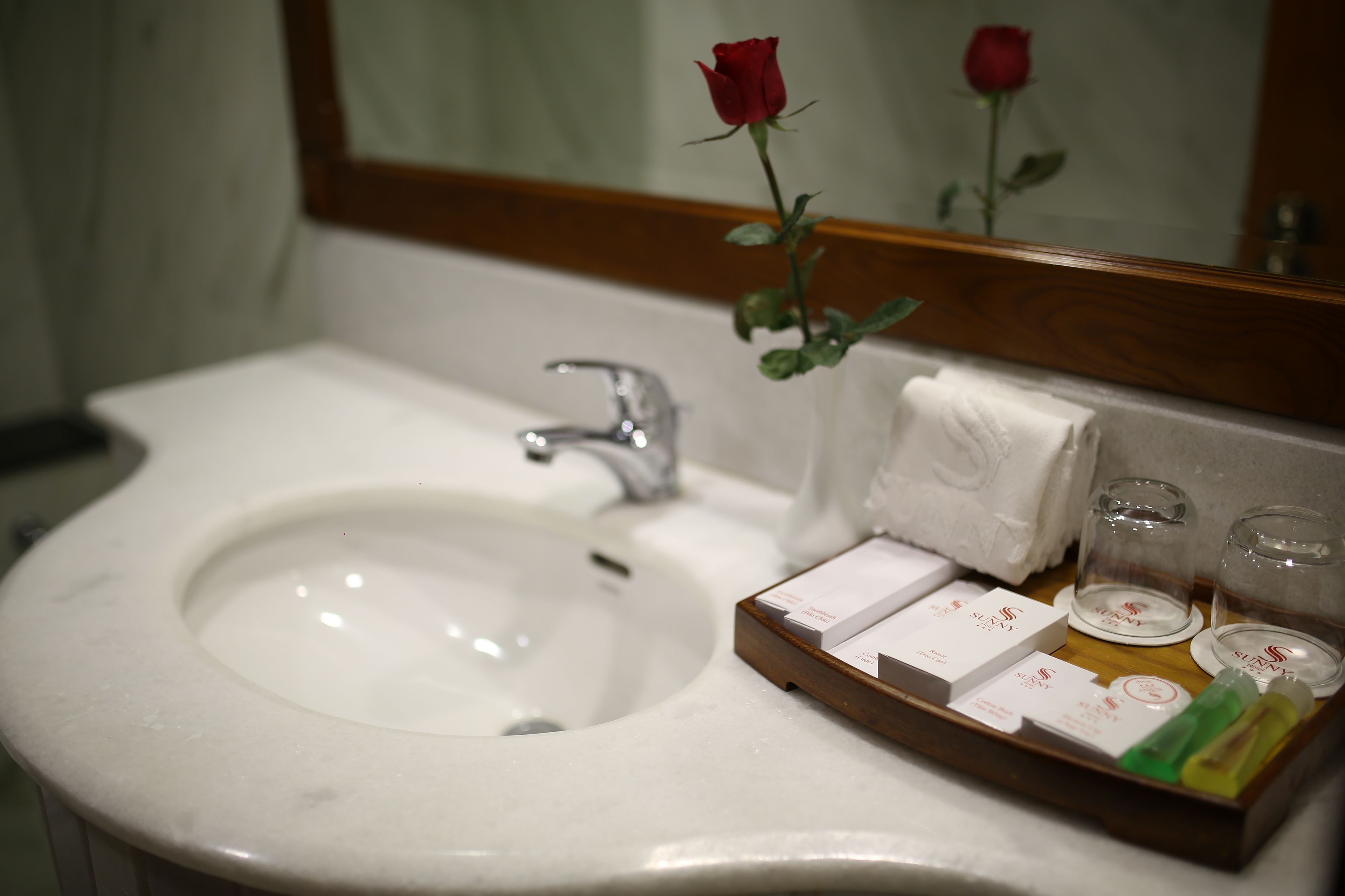 A bathroom sink with a mirror, a rose, and toiletries on a tray.