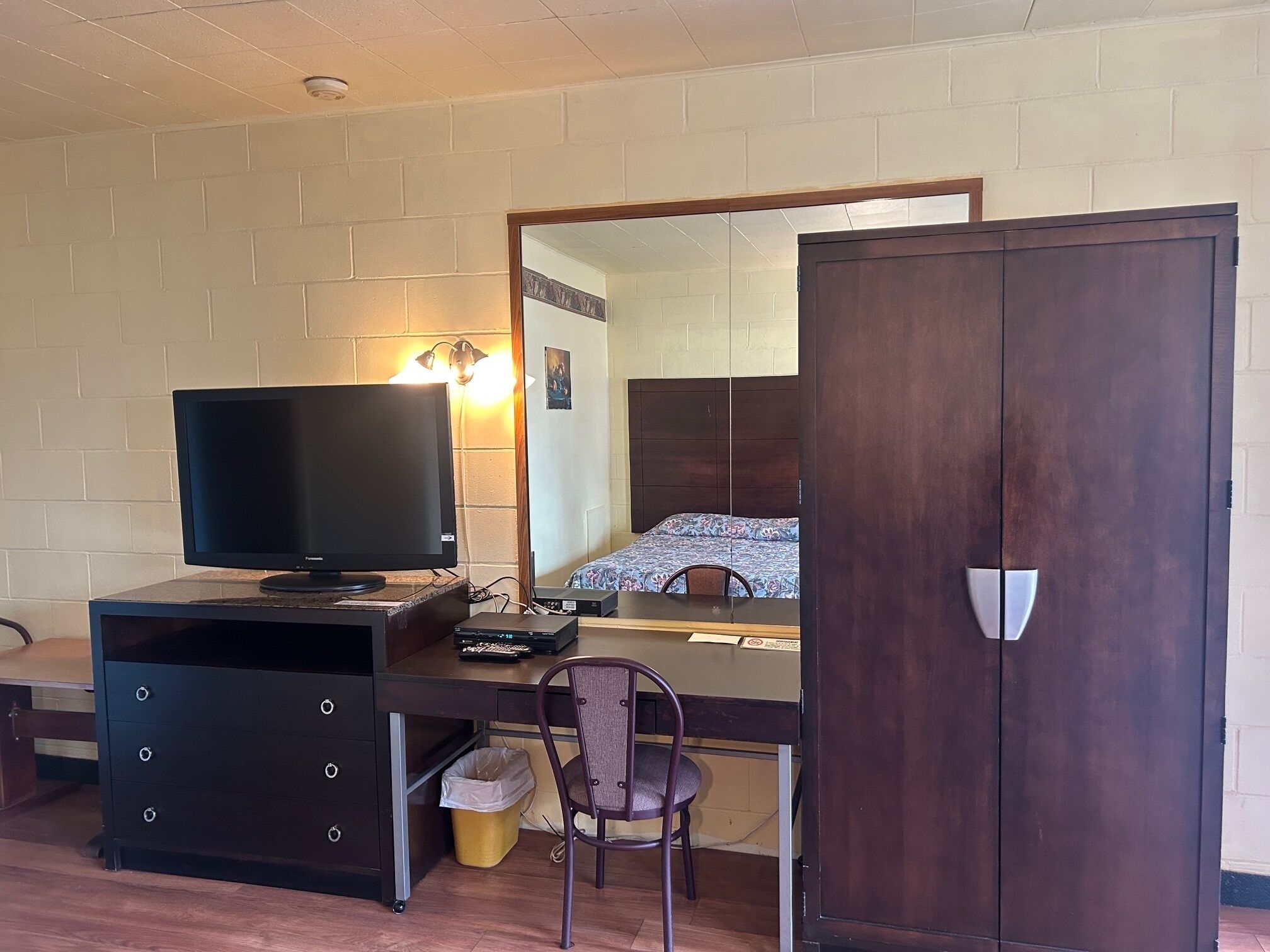 Ein Zimmer mit einem Bett, einem Schreibtisch mit Computer, einem Fernseher, einem Stuhl, einem Kleiderschrank und einer Mülltonne.