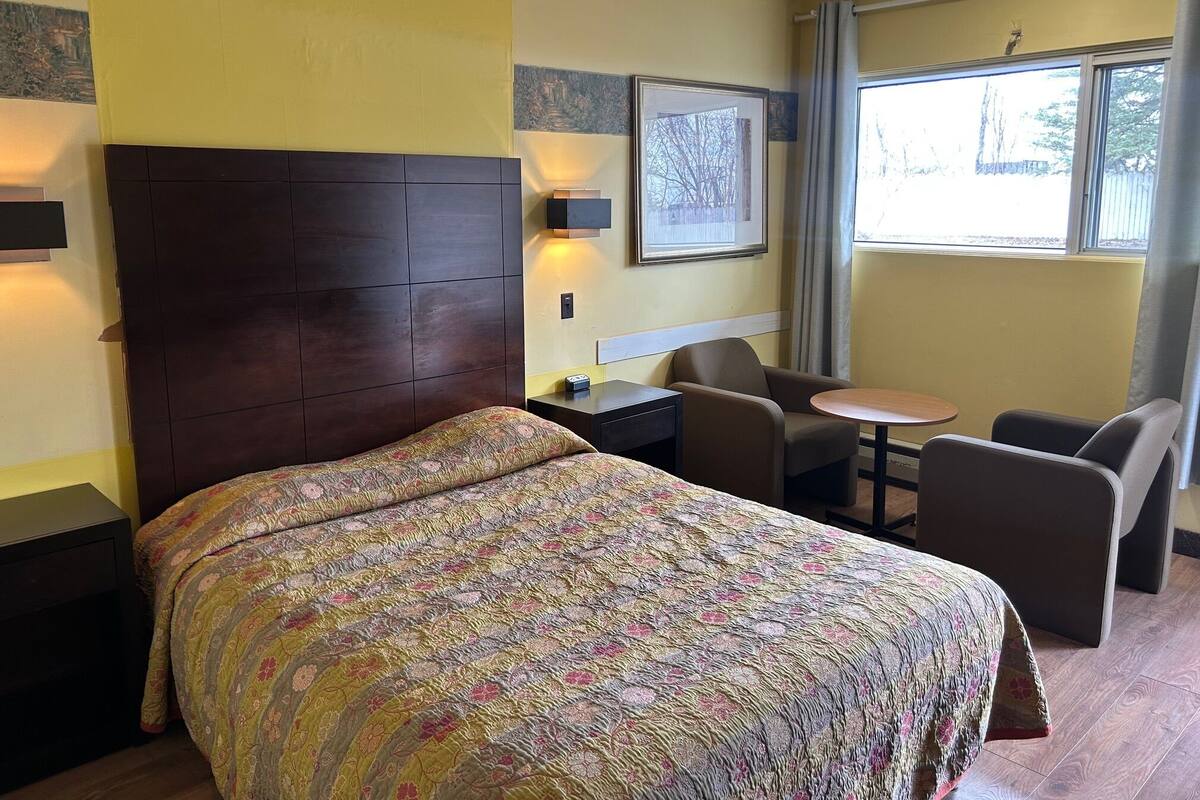 Una habitación de hotel con cama, escritorio, silla, una mesita y ventana con cortinas.
