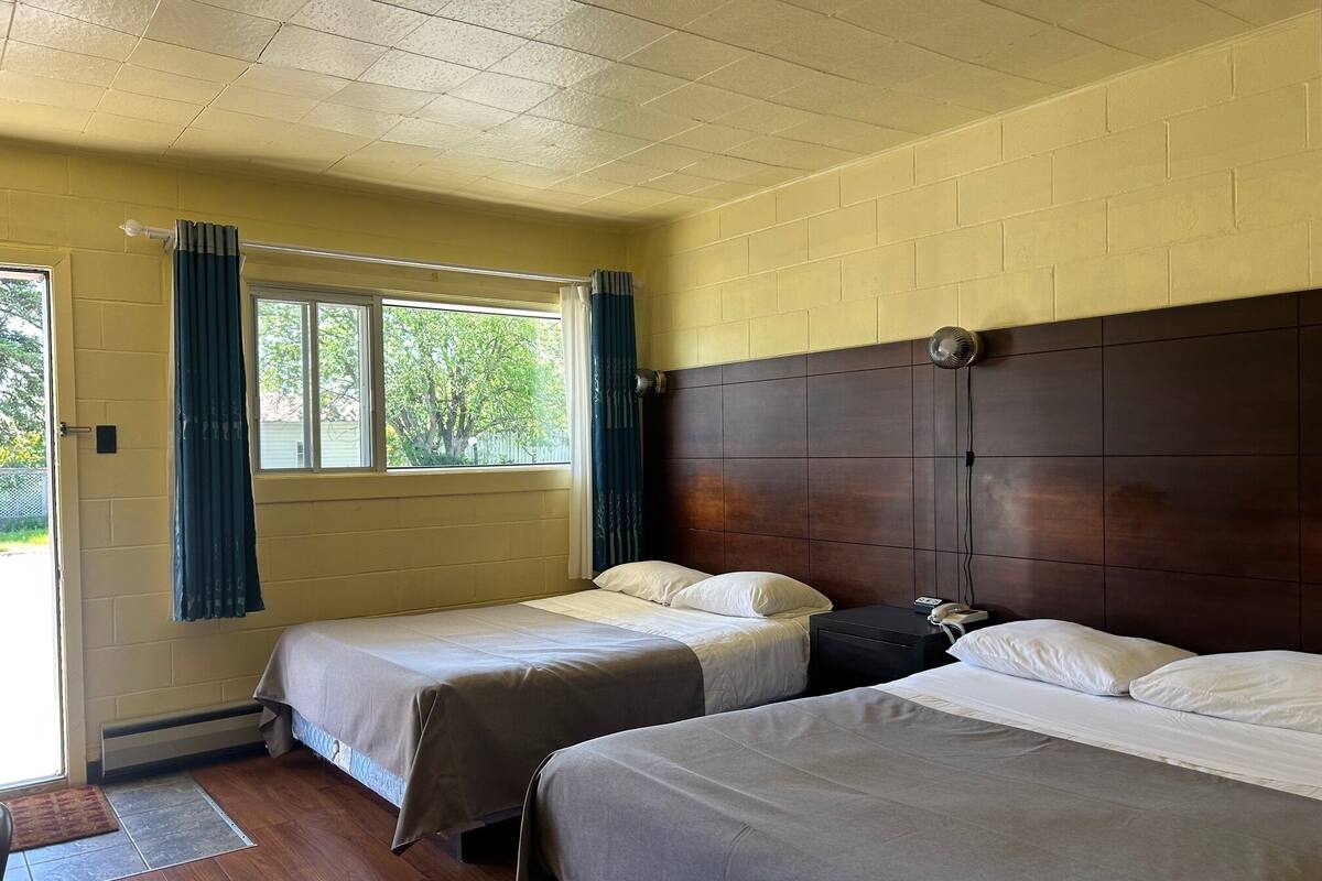 Una habitación con dos camas, piso de madera, pared de ladrillo y una ventana con cortinas.