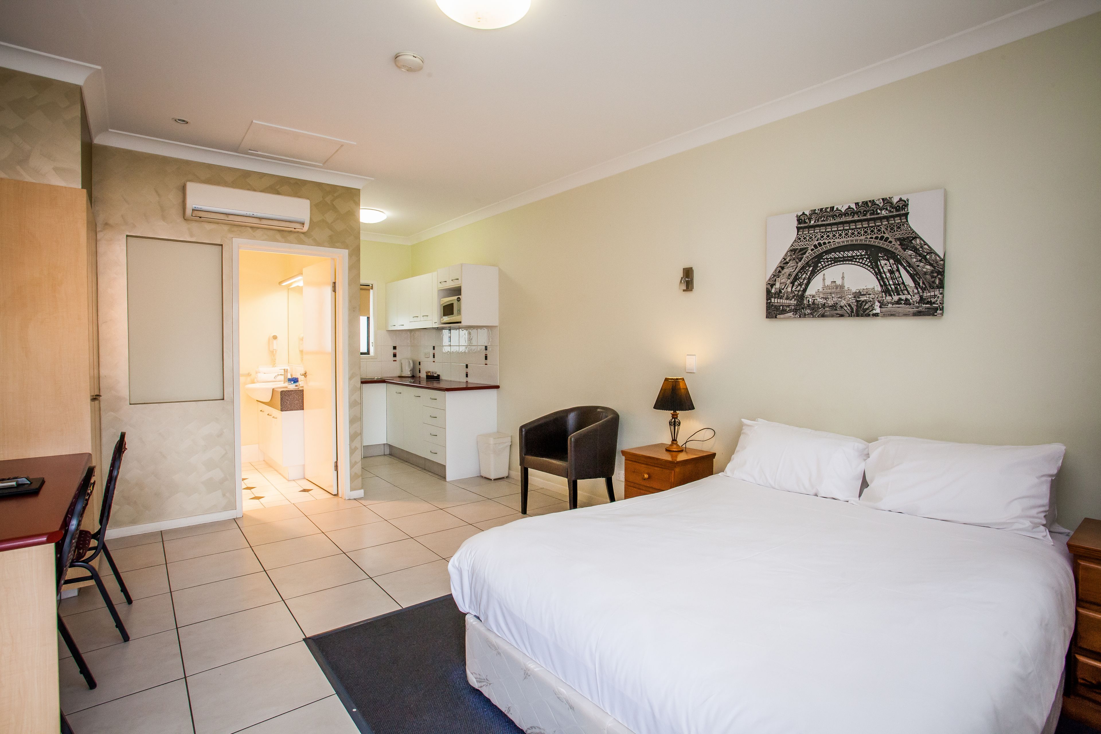 Een hotelkamer met een bed, een bureau, een stoel, een klein tafeltje met een lamp en een foto van de Eiffeltoren aan de muur.