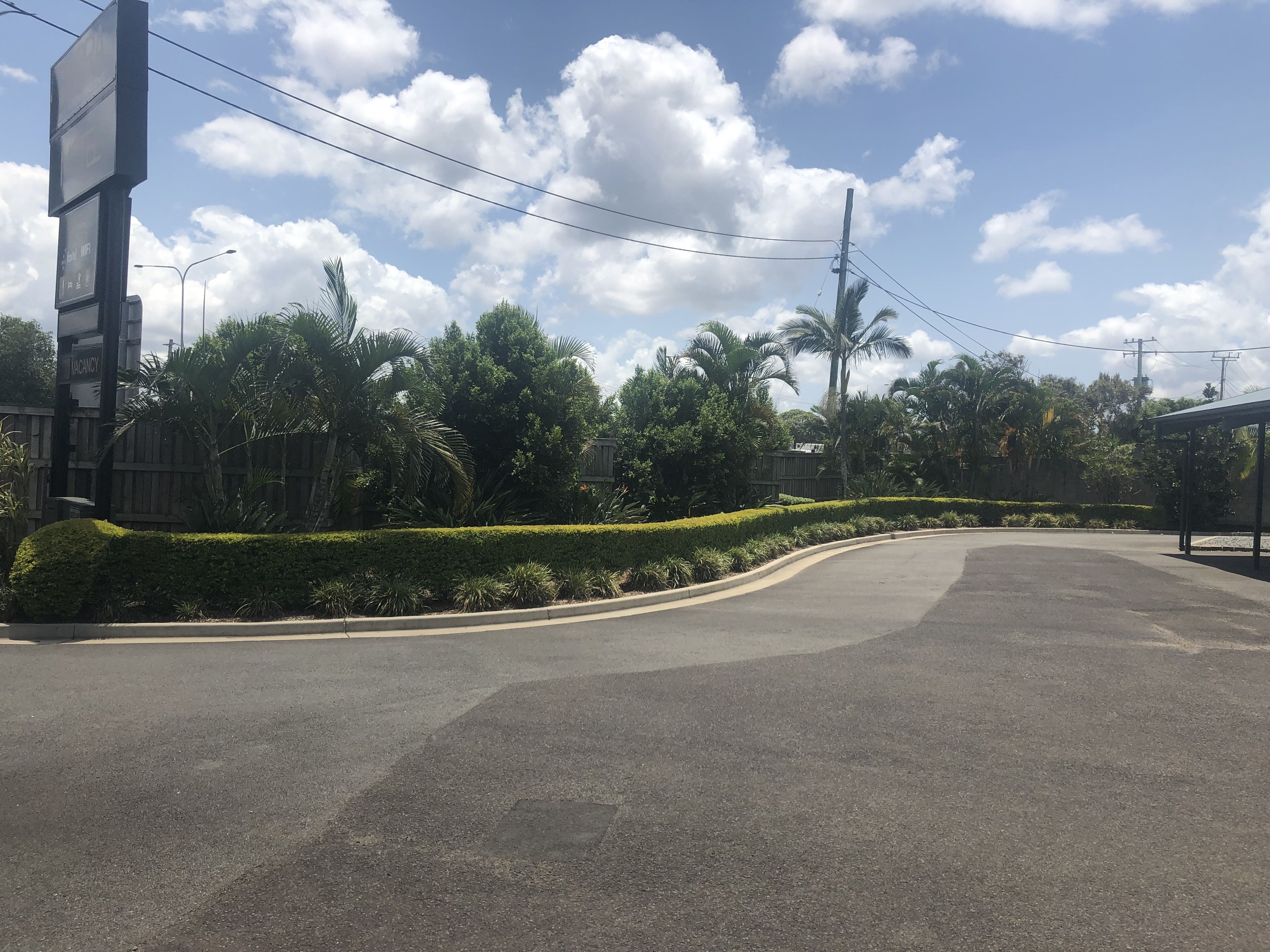 A road with a curved section, surrounded by lush greenery and palm trees. There is a signboard on the left side, and a building with a signboard on the right.
