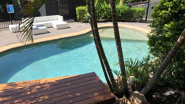 A clear swimming pool with a wooden deck in the foreground, surrounded by lush greenery and a white outdoor seating area. - Darra Motel and Conference Centre (Darra)