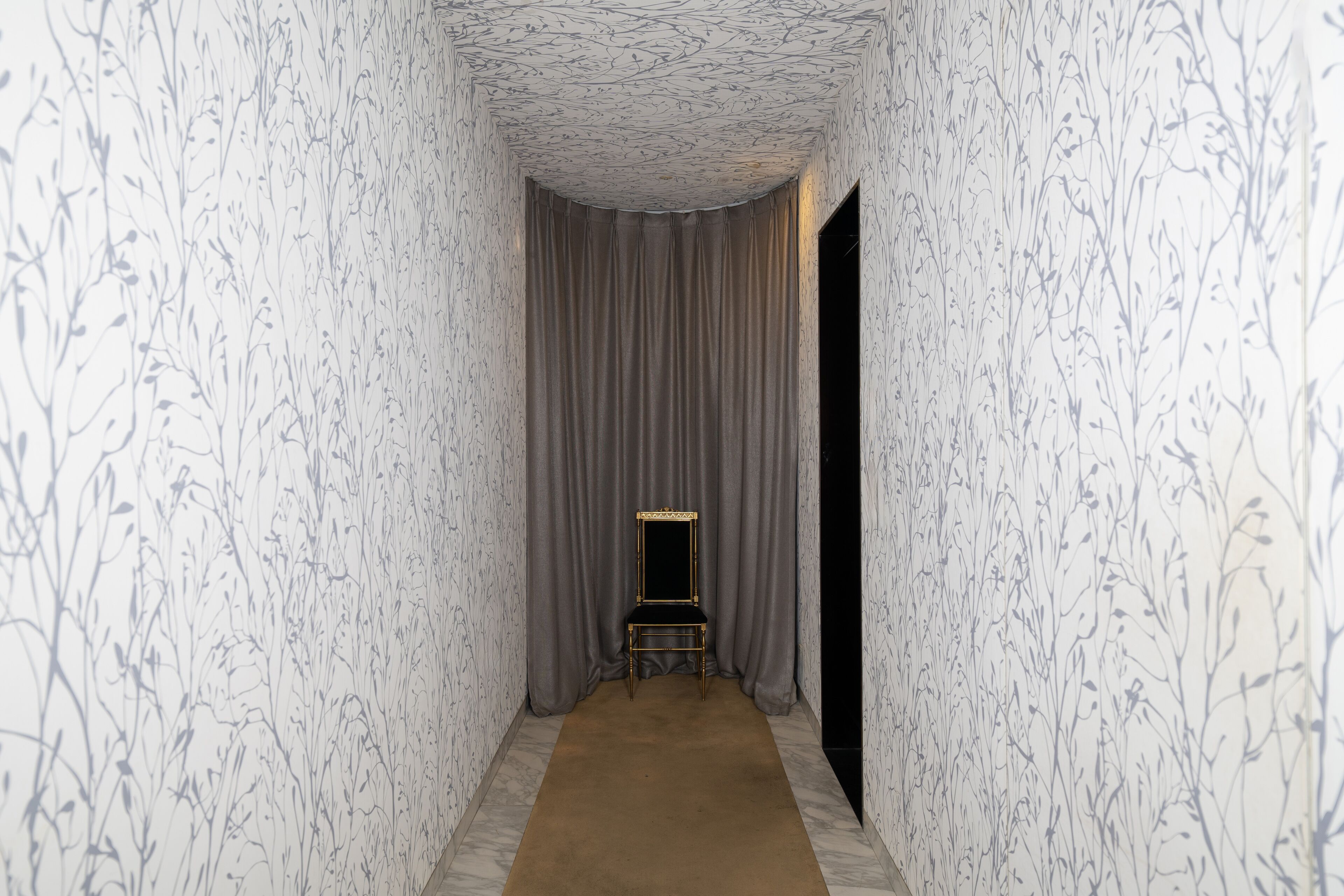 A narrow hallway with patterned wallpaper, a dark curtain, and a small chair with a mirror.