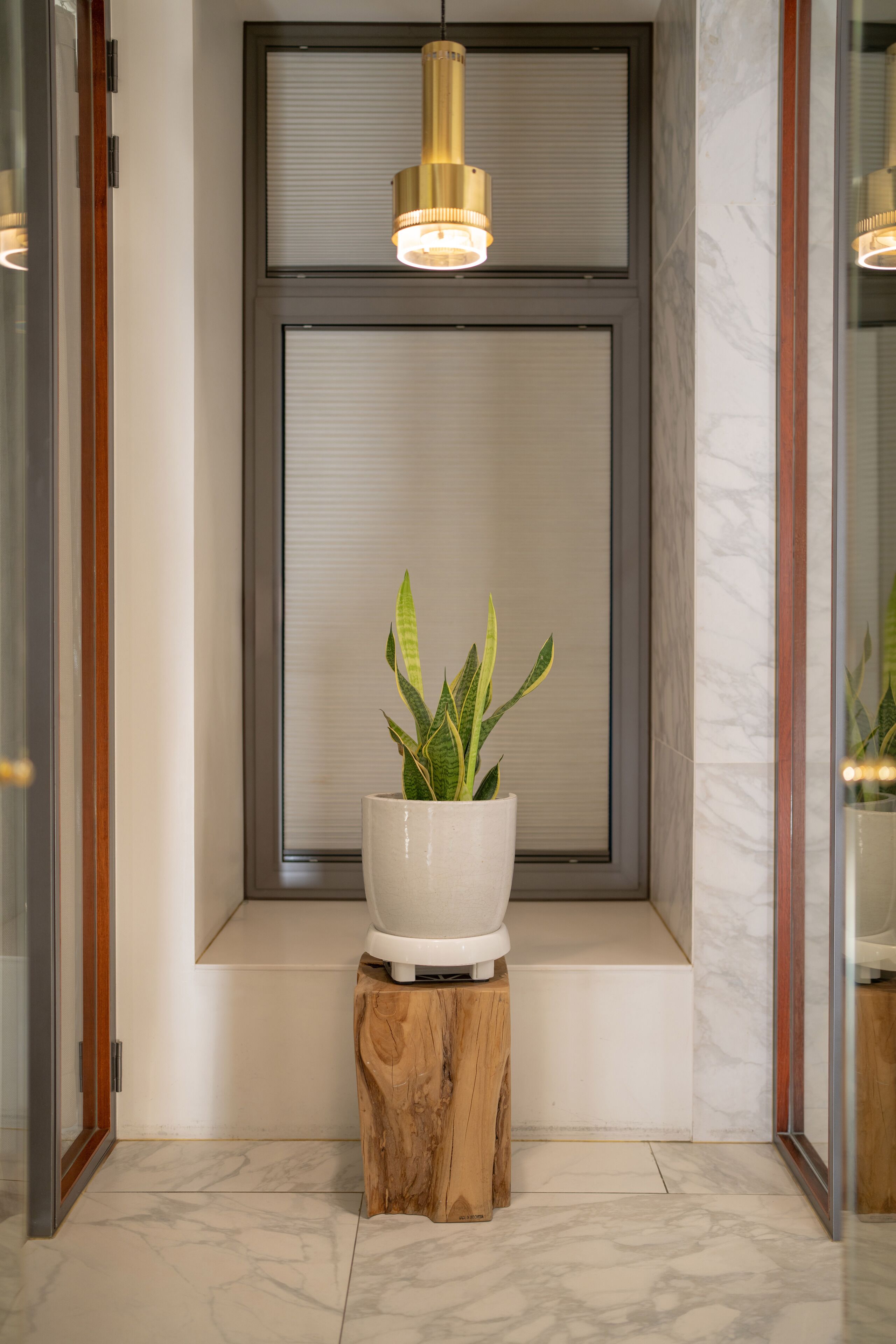 Un interior moderno con una planta en maceta sobre un taburete de madera, un ventanal con persianas y una lámpara colgante.
