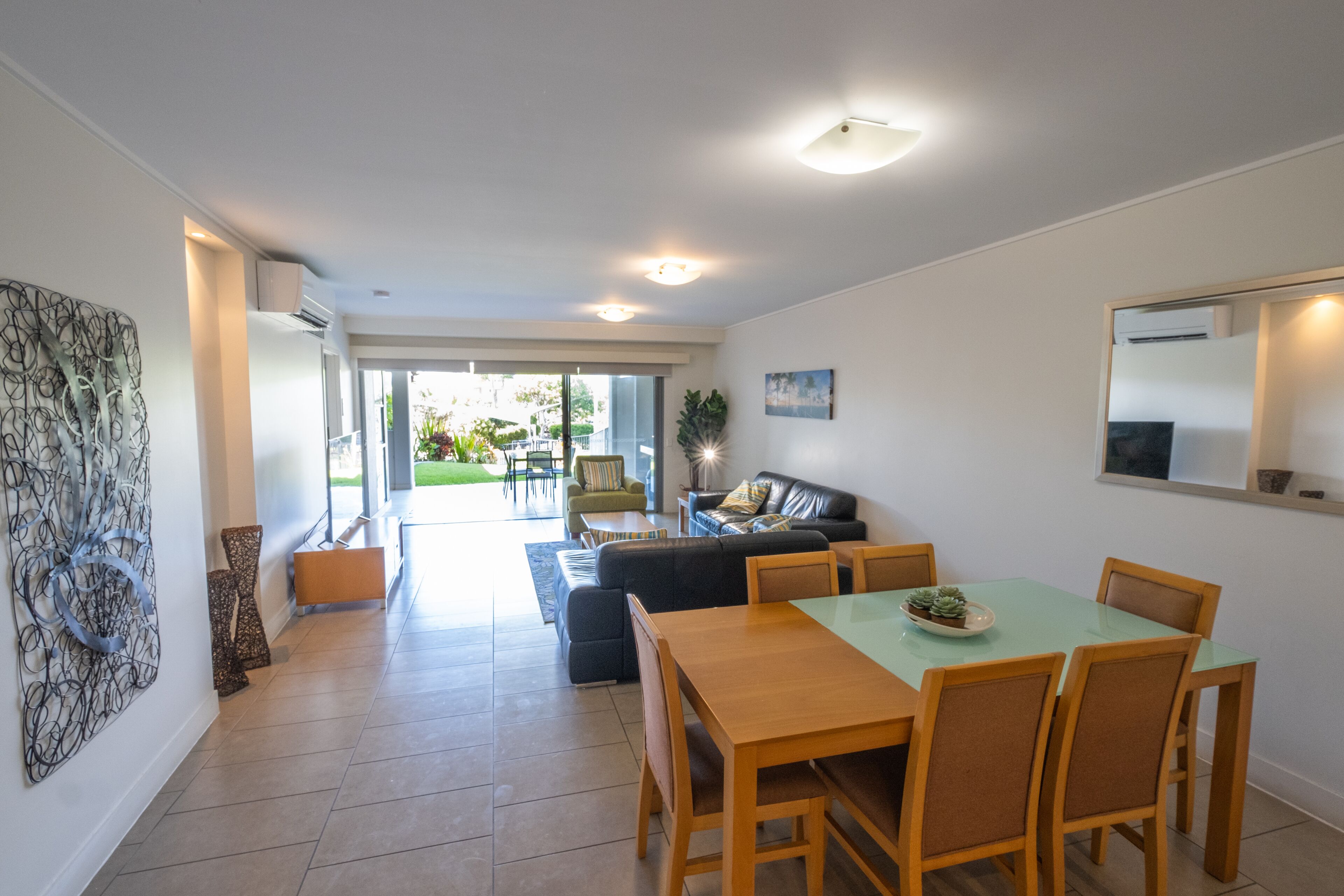 A modern living room with a sofa, coffee table, and dining table with chairs. There is a large window with a view of the outdoors.