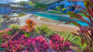 A pool area with a sandy beach, surrounded by lush greenery and a covered seating area. - Pavillions on 1770 (Agnes Water)