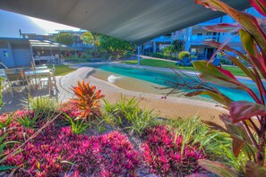 A pool area with a sandy beach, surrounded by lush greenery and a covered seating area. - Pavillions on 1770 (Agnes Water)