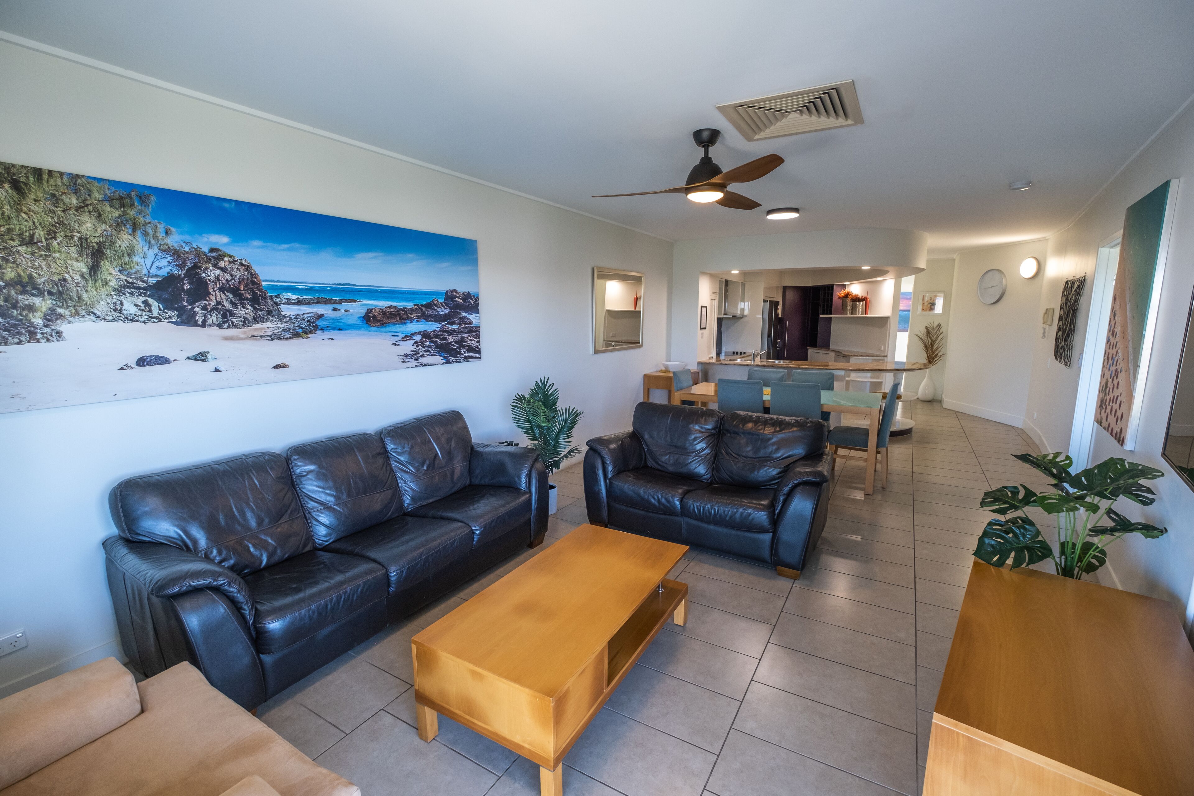 A modern living room with leather sofas, a wooden coffee table, and a large beach-themed painting on the wall.
