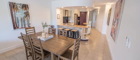 A dining area with a wooden table and chairs, a kitchen in the background, and artwork on the walls.