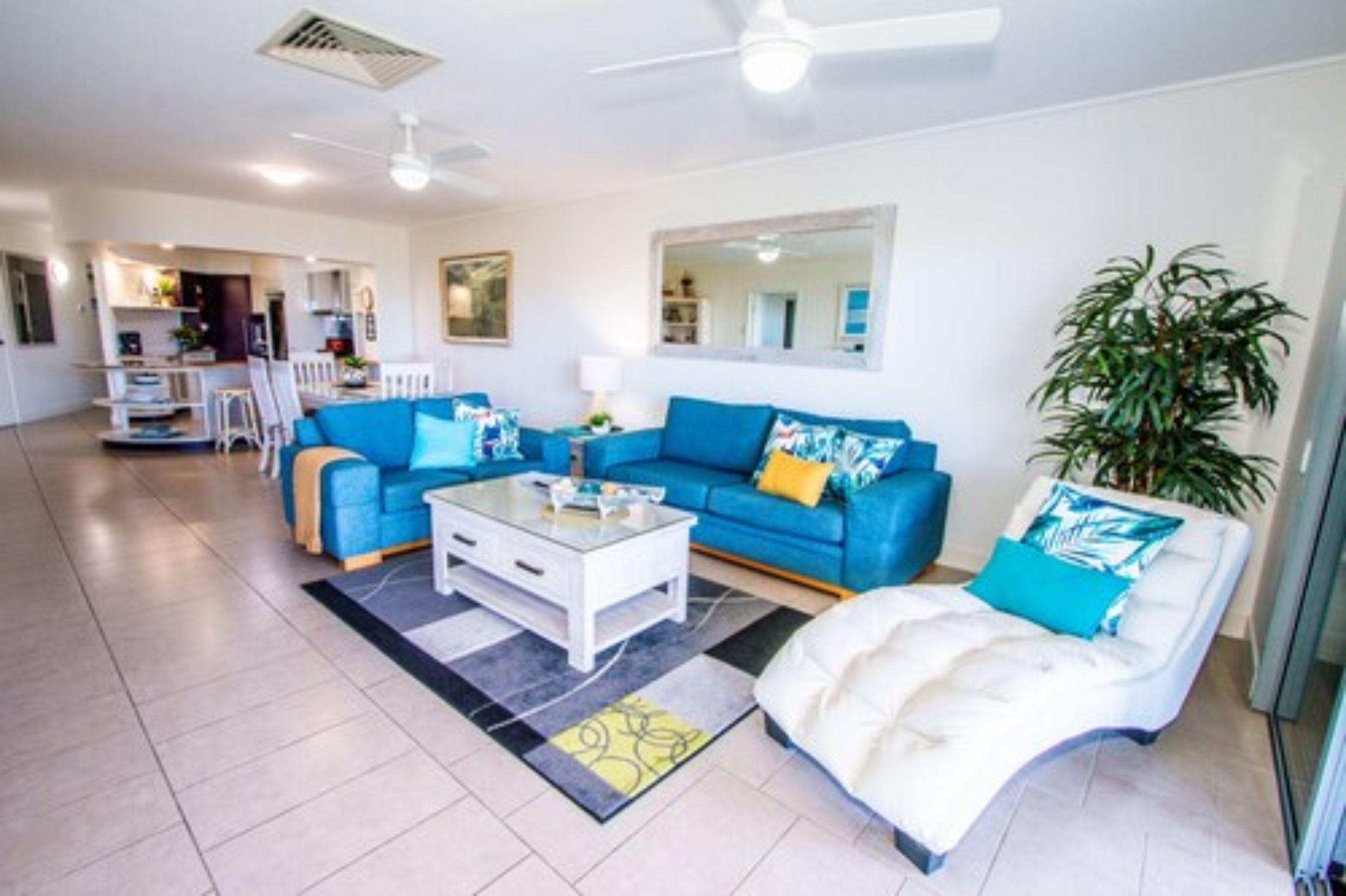 A living room with a blue sofa, a white armchair, and a coffee table. There is a dining area in the background.