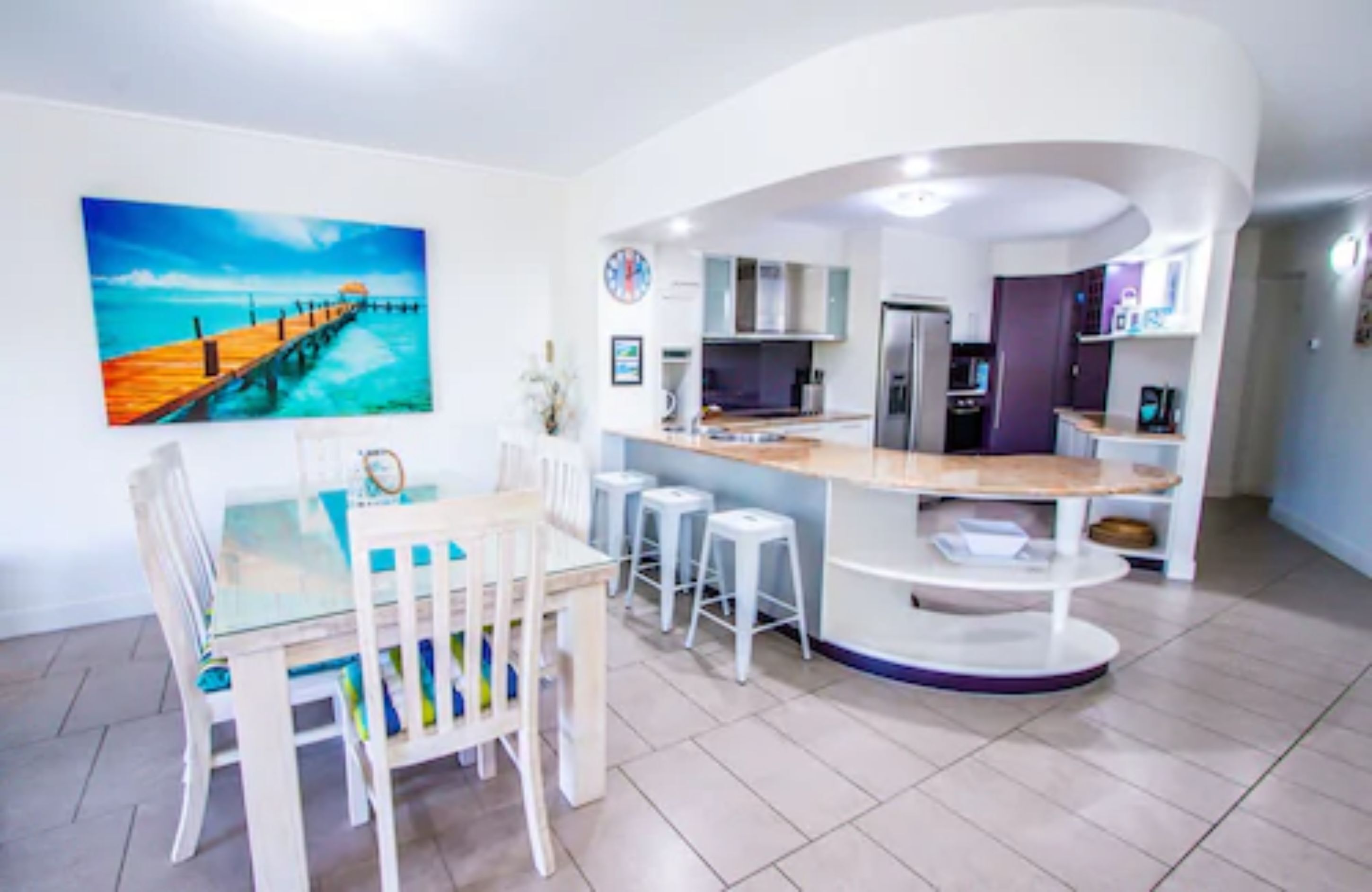 A modern kitchen with a central island, bar stools, and a dining area with a glass table and chairs.