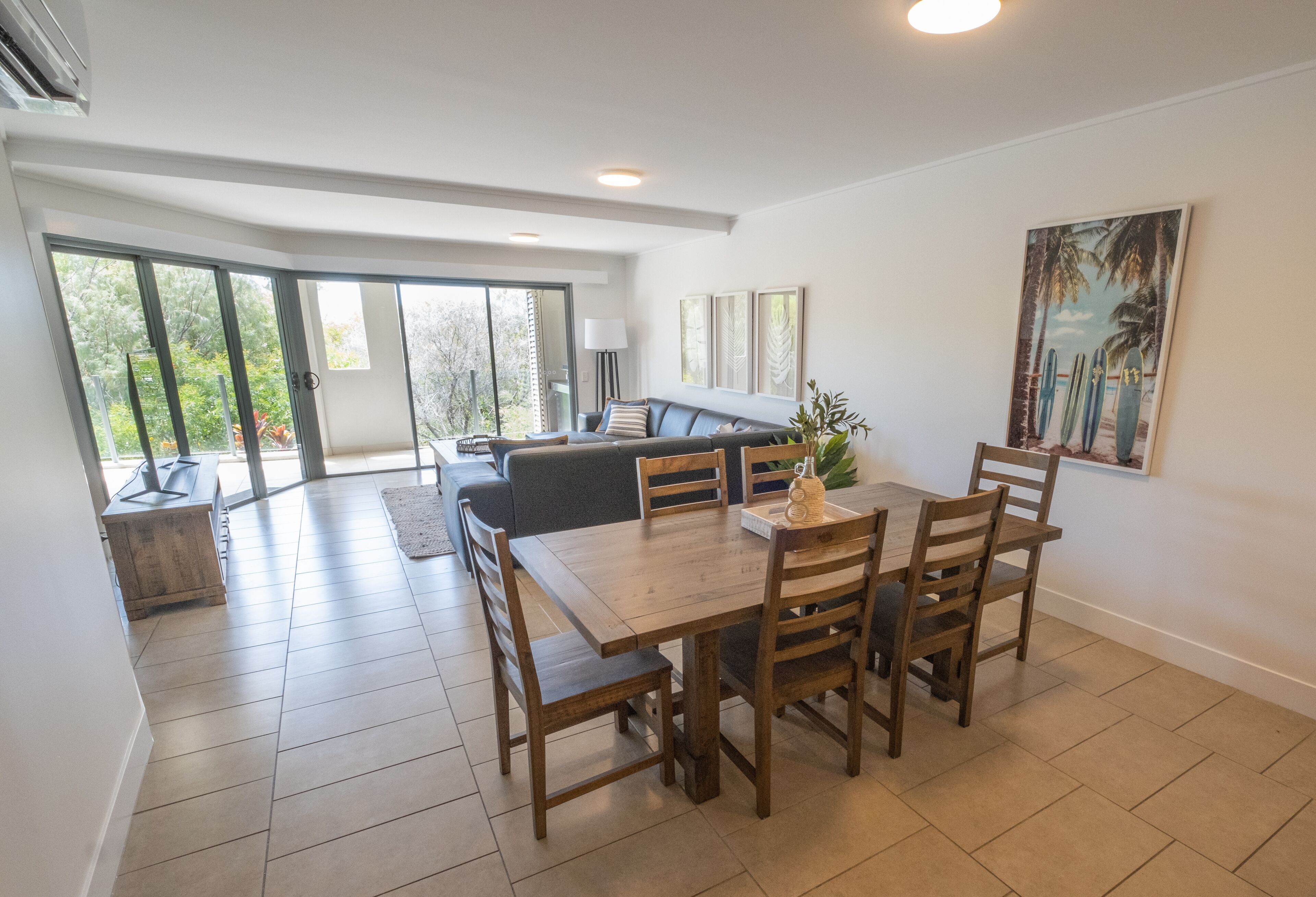 A spacious dining area with a wooden table and chairs, a sofa, and a large window with a view of greenery.