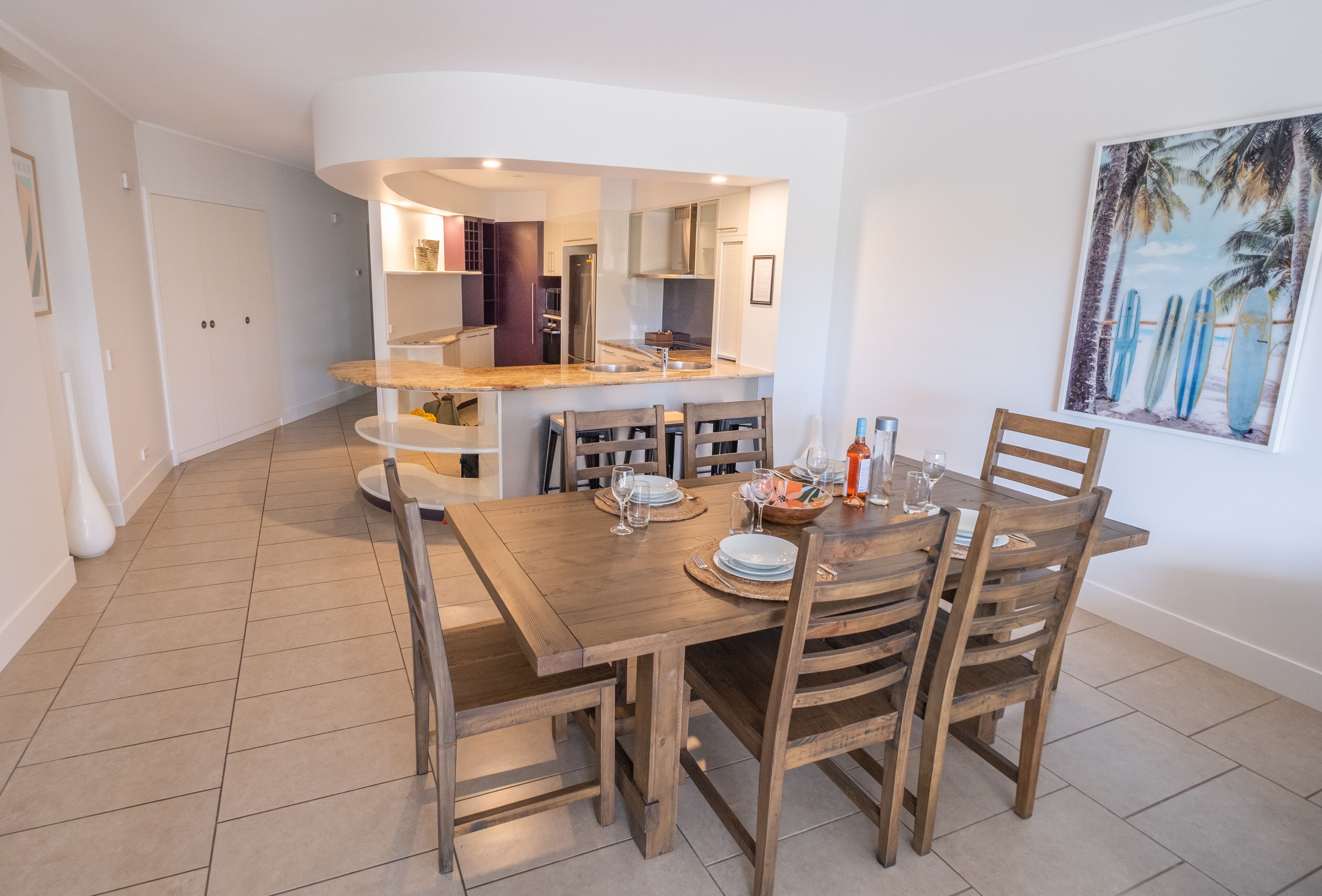 A dining area with a wooden table and chairs, a kitchen in the background, and a beach-themed painting on the wall.