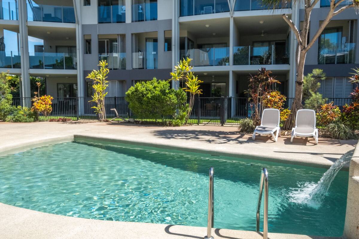 A swimming pool with a clear blue water, surrounded by a concrete deck, and two white lounge chairs.