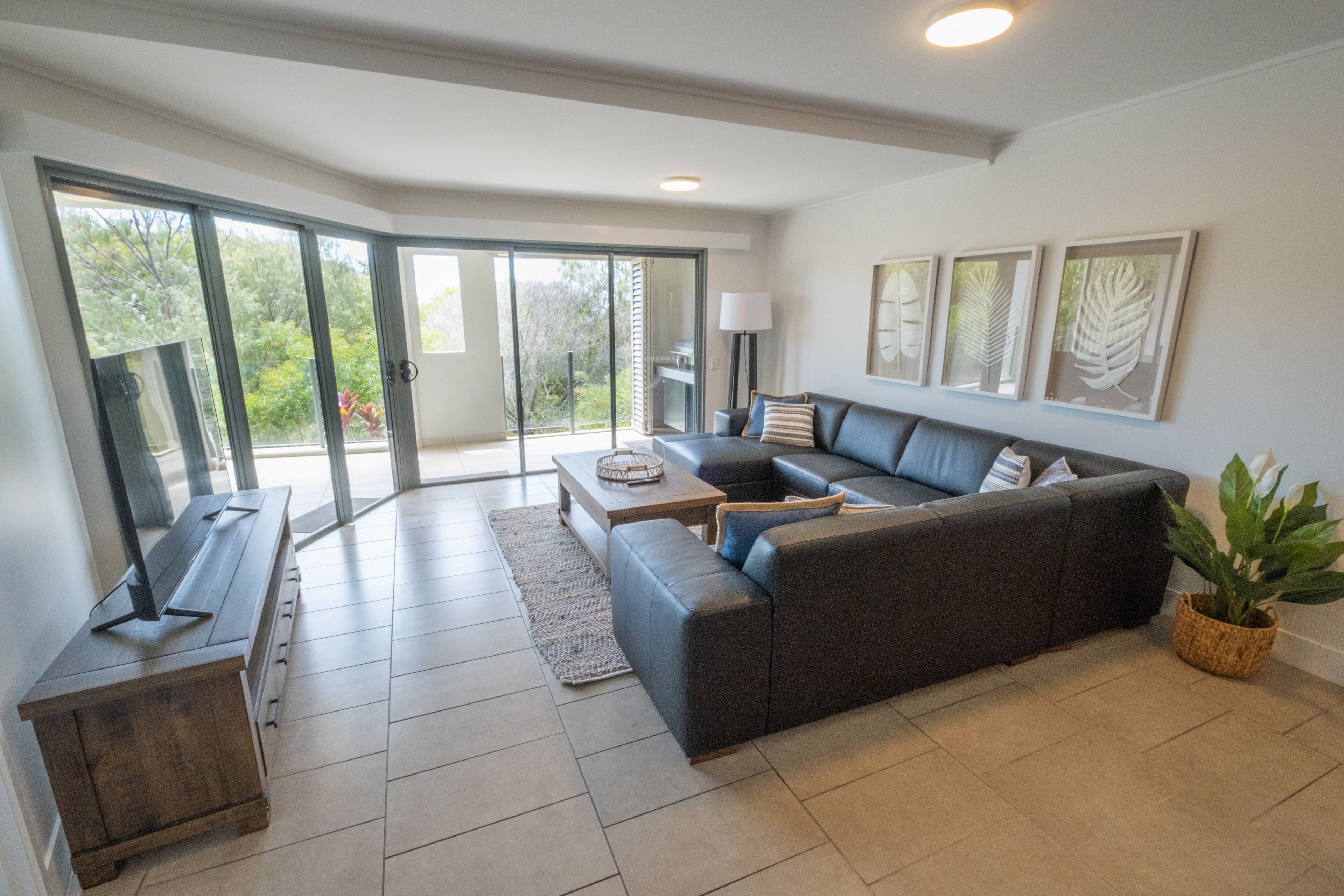 Ein modernes Wohnzimmer mit einem dunkelgrauen Sofa, einem Holztisch und einer TV-Konsole. Eine Glastür öffnet sich zu einem Balkon mit Blick auf Bäume.
