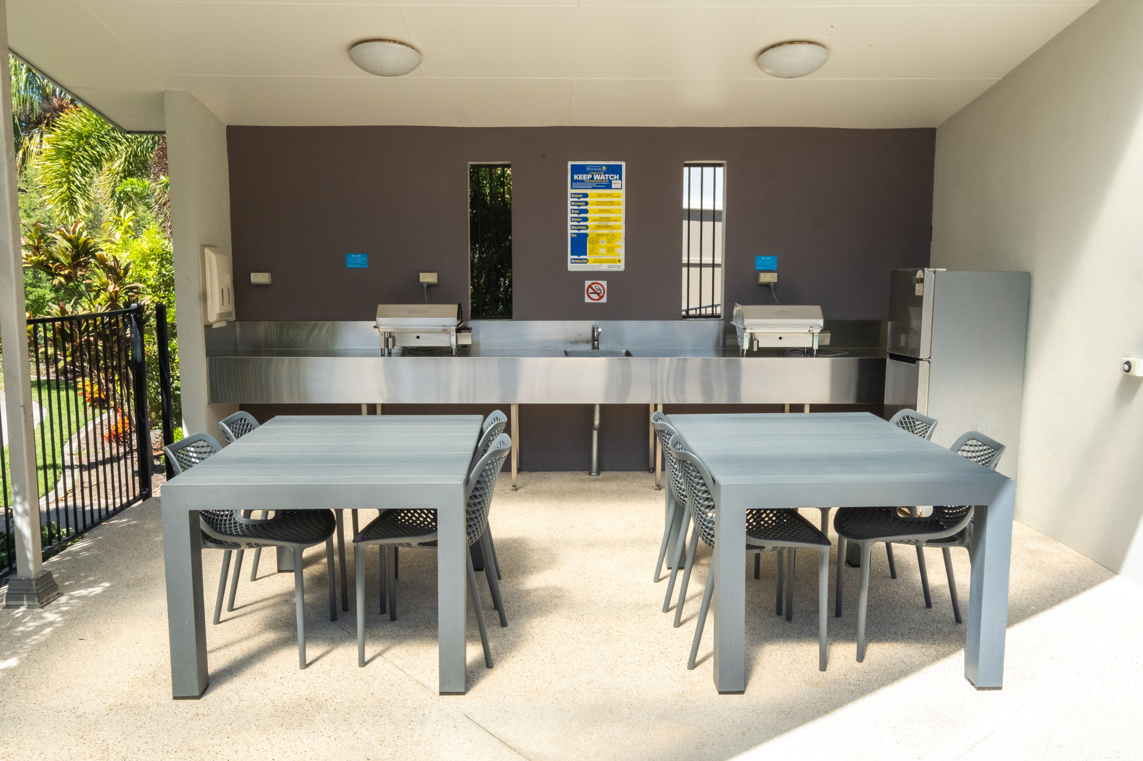A modern outdoor kitchen with a grey table and chairs, a stainless steel refrigerator, and a 'Keep Watch' sign on the wall.