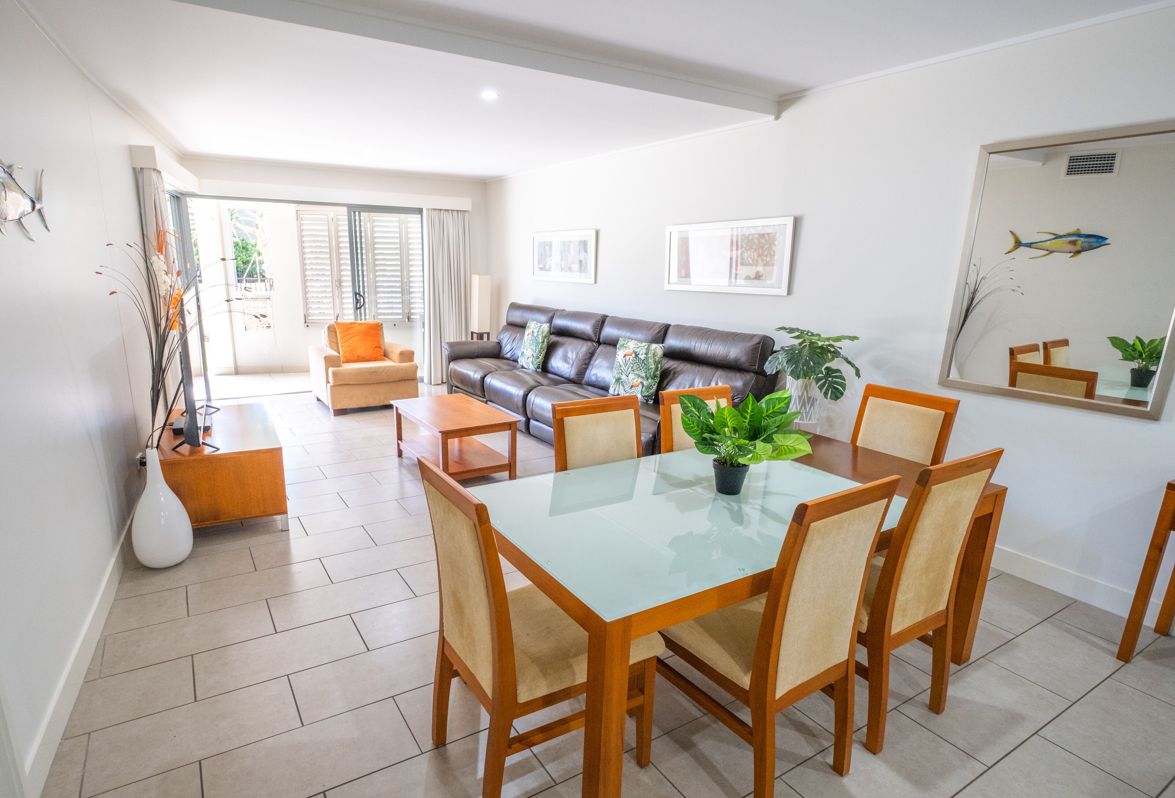A dining area with a glass table and wooden chairs, a leather sofa, and a large mirror on the wall.