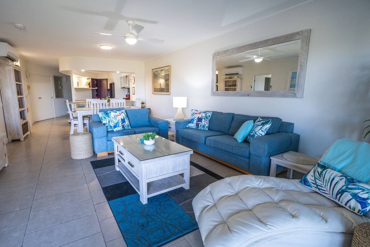 A living room with a blue sofa, a white sofa, a coffee table, and a large mirror on the wall.