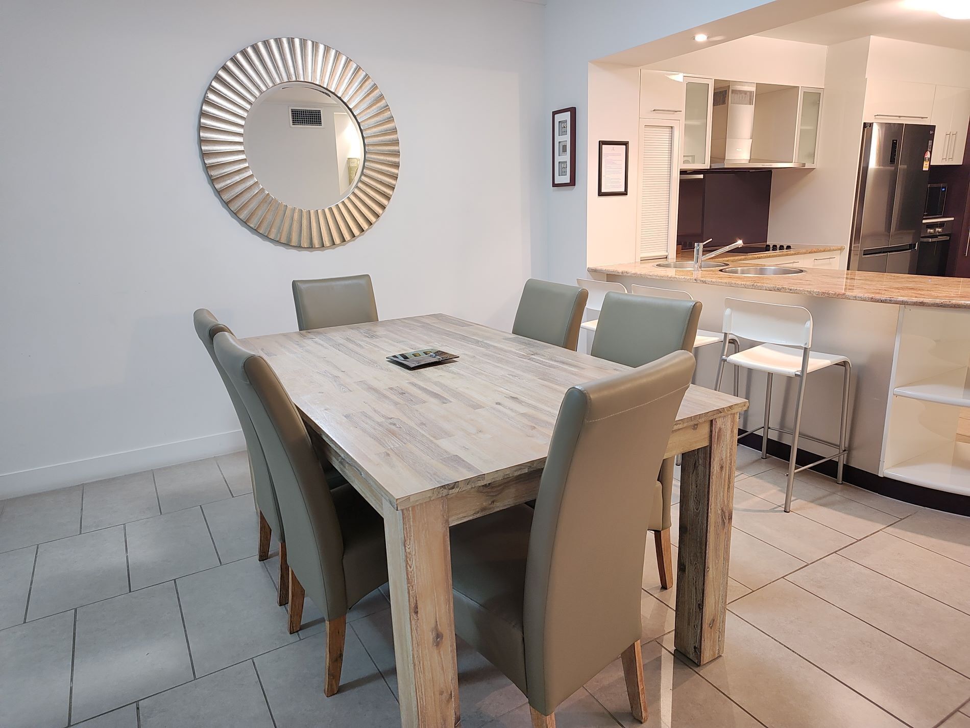 Un comedor con una mesa de madera y seis sillas, un espejo redondo en la pared, y una zona de cocina con electrodomésticos modernos.