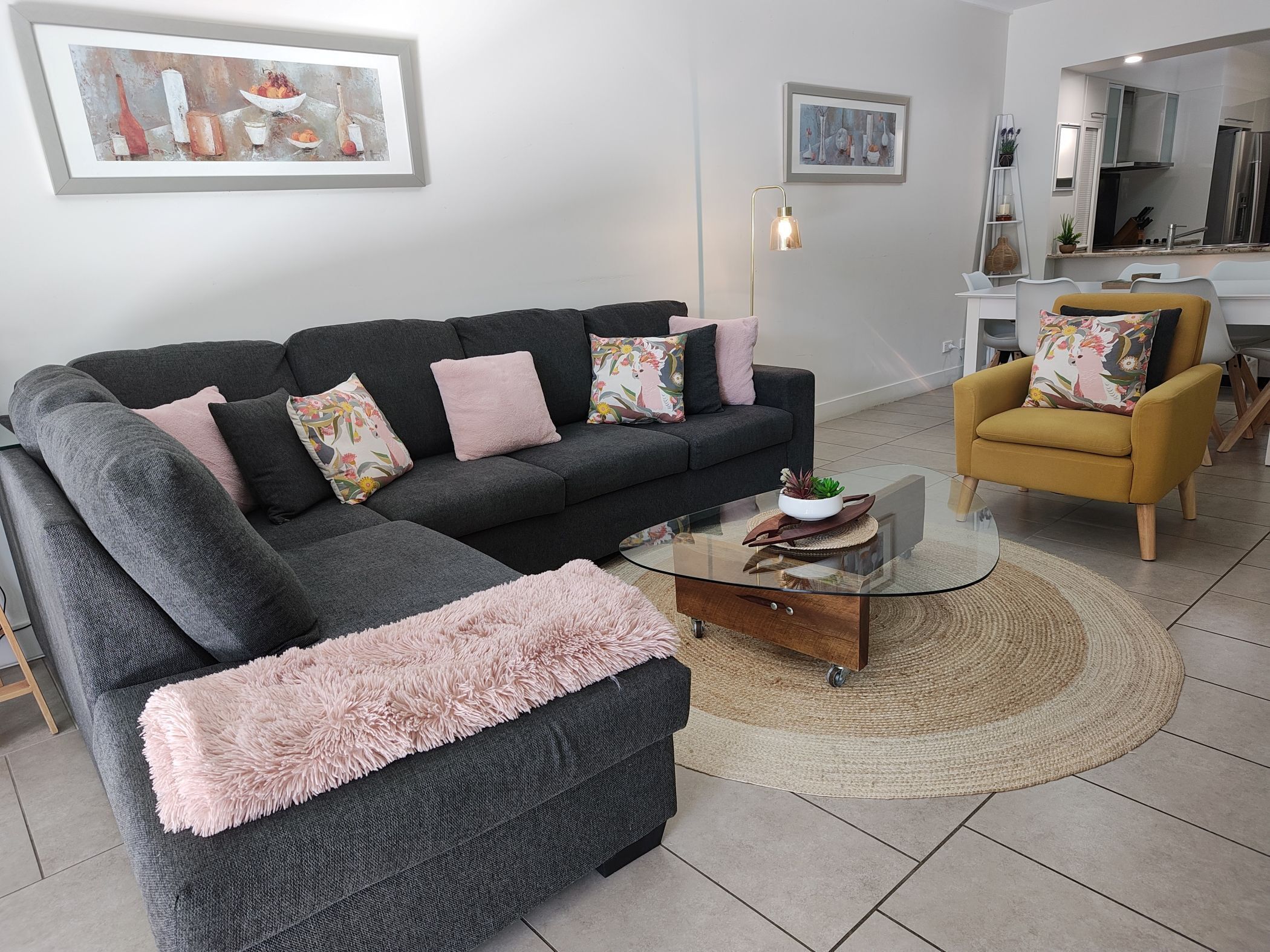 Una sala moderna con un sofá seccional gris, una mesa de centro de vidrio, un sillón amarillo y una zona de cocina al fondo.