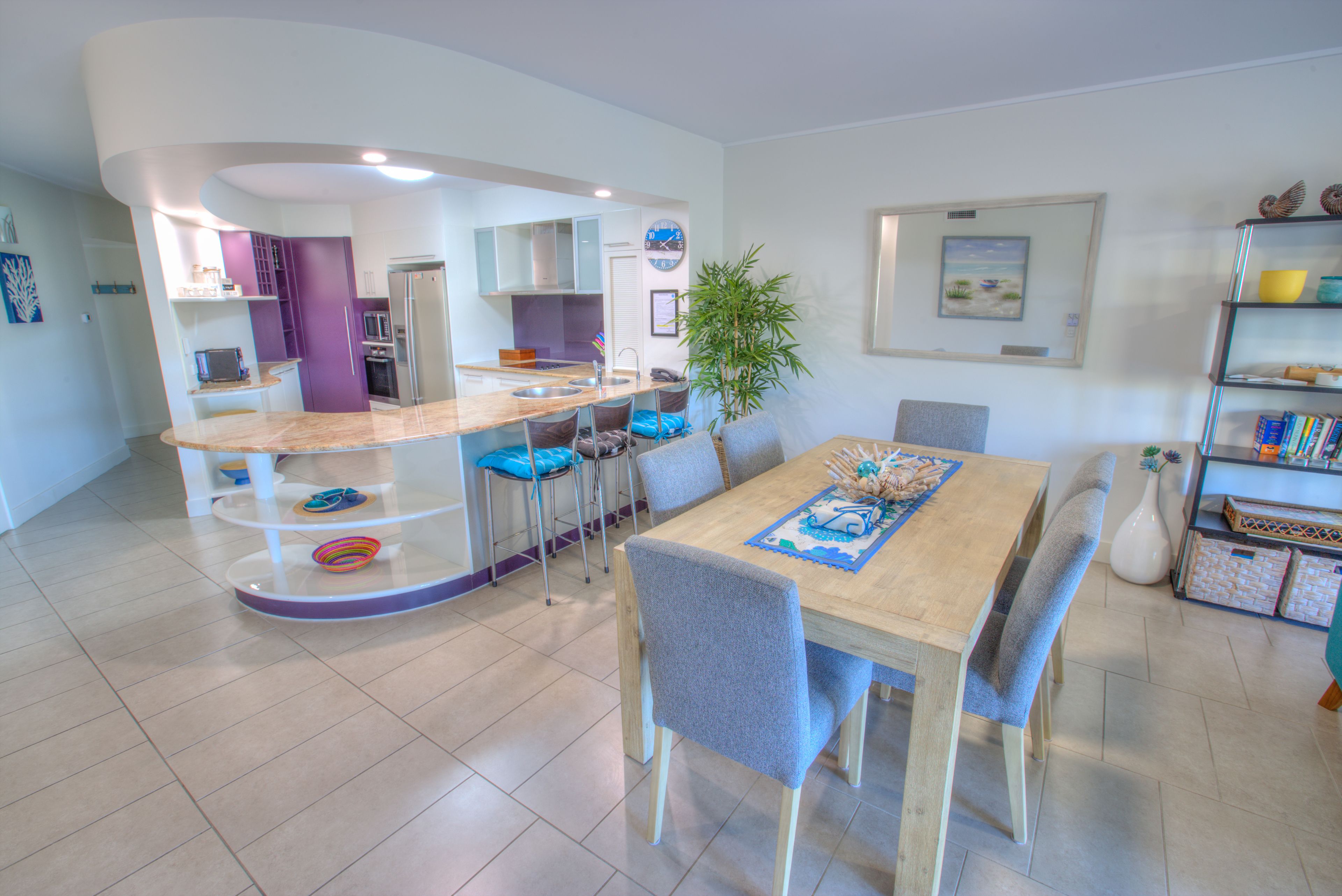 A modern kitchen with a curved island, a dining area with a wooden table and chairs, and a bookshelf with decorative items.