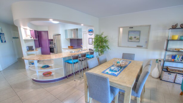 A modern kitchen with a curved island, a dining area with a wooden table and chairs, and a bookshelf with decorative items. - Pavillions on 1770 (Agnes Water)