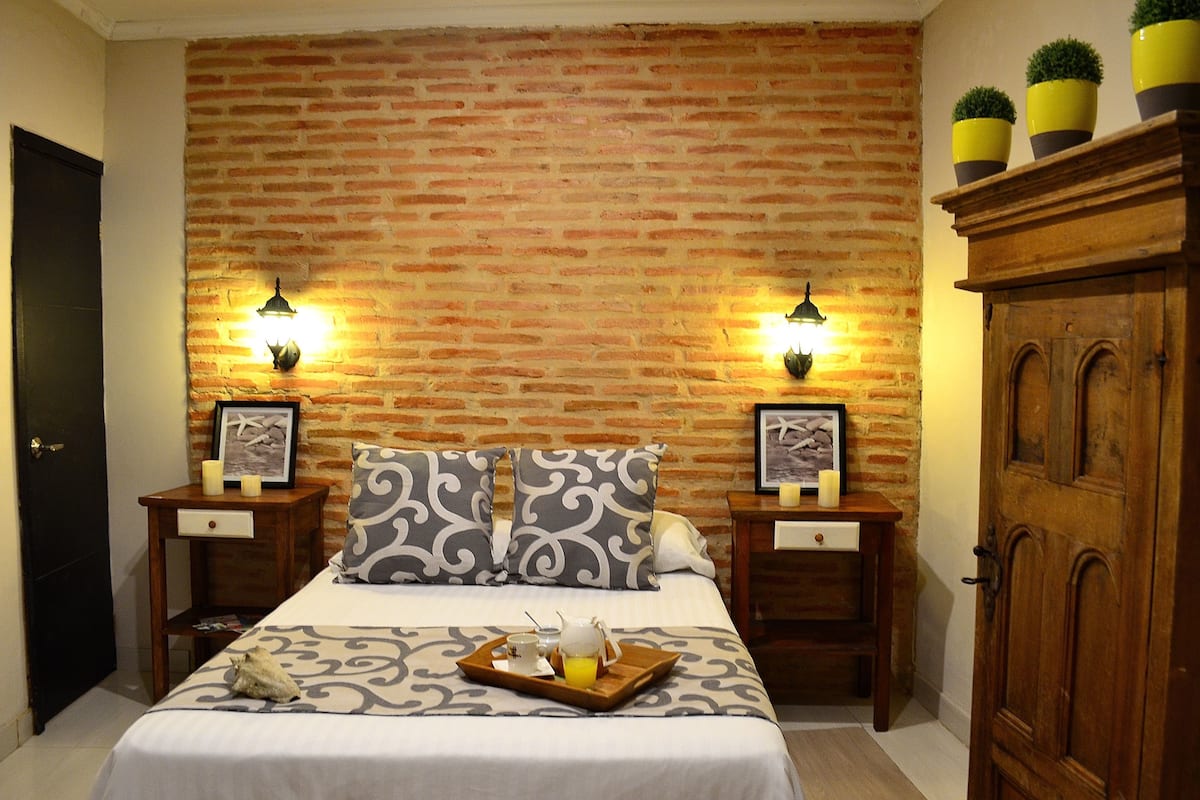 A hotel room with a brick wall, two bedside tables with lamps, a bed with patterned bedding, a tray with breakfast items, and a wooden wardrobe.
