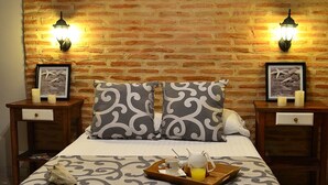 A neatly made bed with a tray of breakfast items, two bedside tables with lamps, and a brick wall backdrop. - Hotel Galería la Trinidad (Cartagena)
