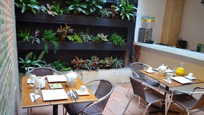 A dining area with a wooden table set for four, surrounded by wicker chairs. A vertical garden with various plants is behind the table. - Hotel Galería la Trinidad (Cartagena)