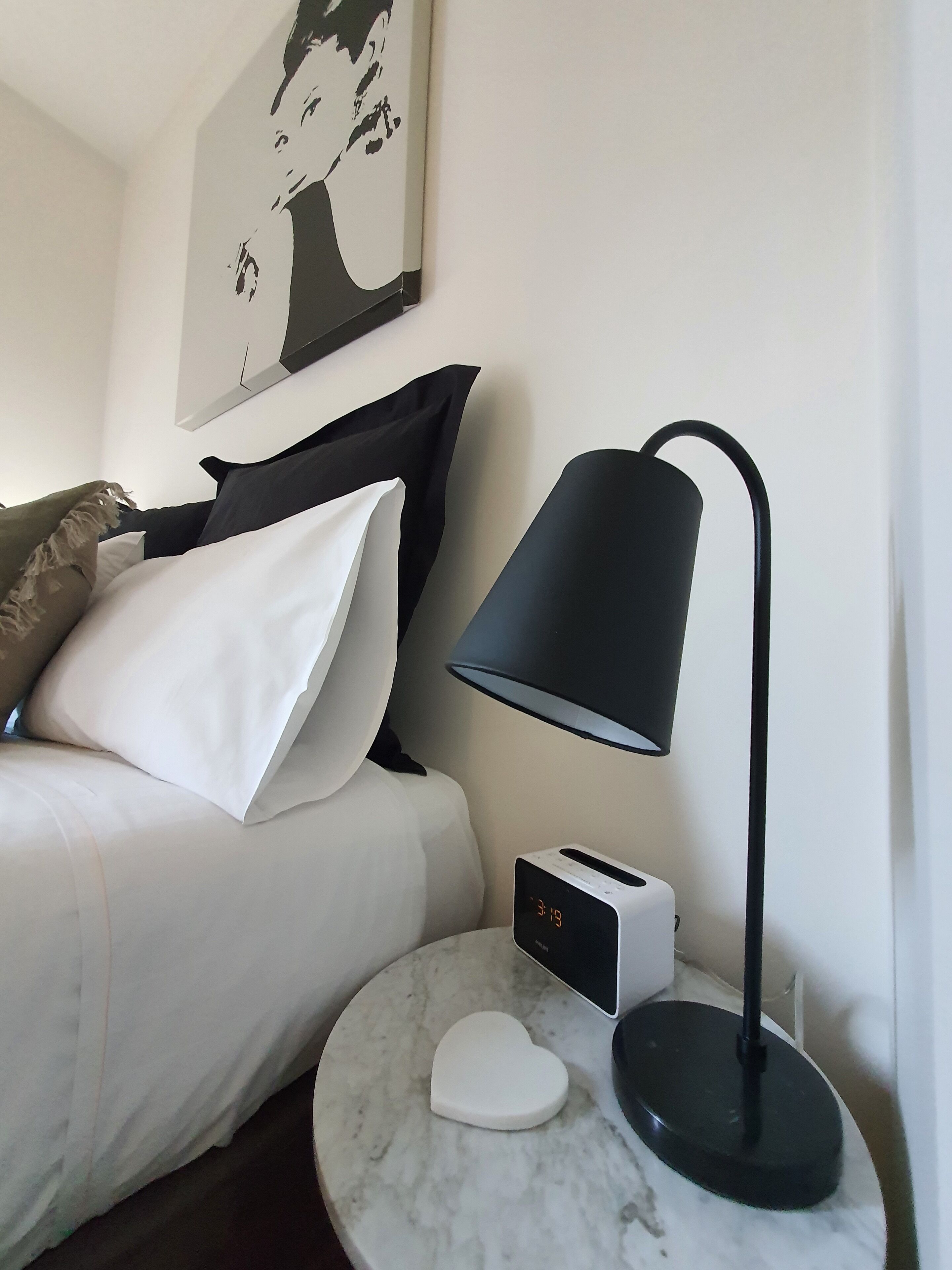 A neatly made bed with white and black bedding, a modern black desk lamp, a digital clock, and a heart-shaped object on a marble table.