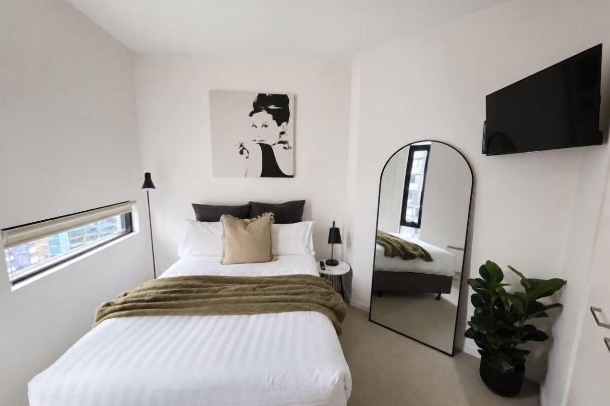 Une chambre à coucher avec un lit, une table de chevet, un miroir, une télévision, une fenêtre et une plante.