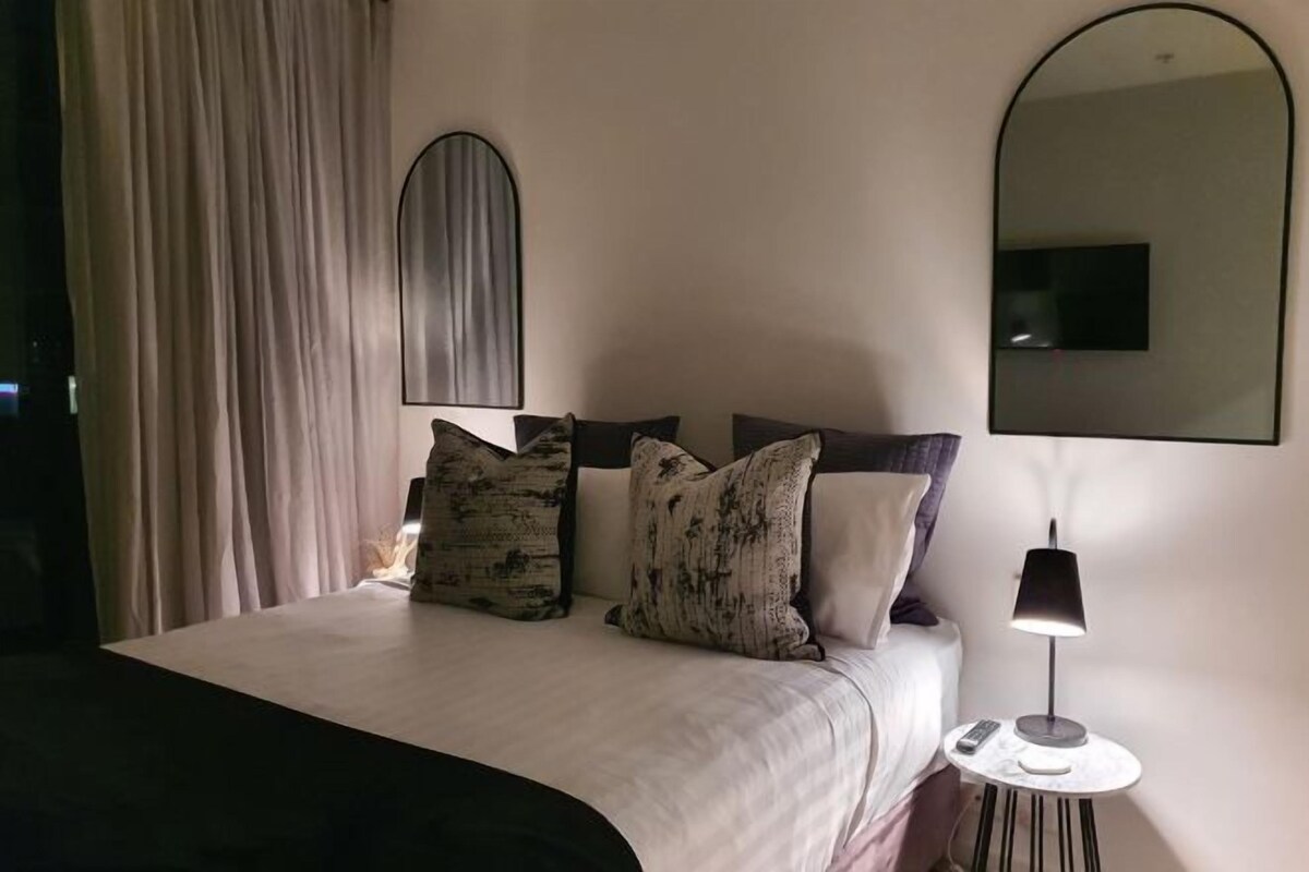 A neatly made bed with a white comforter and several patterned pillows, a bedside lamp, and a small table with a remote control.