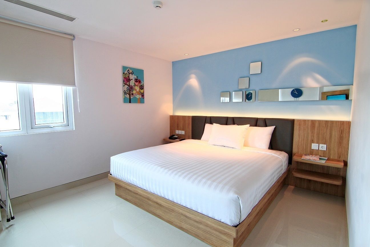 Une chambre d’hôtel moderne avec un grand lit, des tables de chevet, une petite table, une horloge murale et une fenêtre avec des stores.