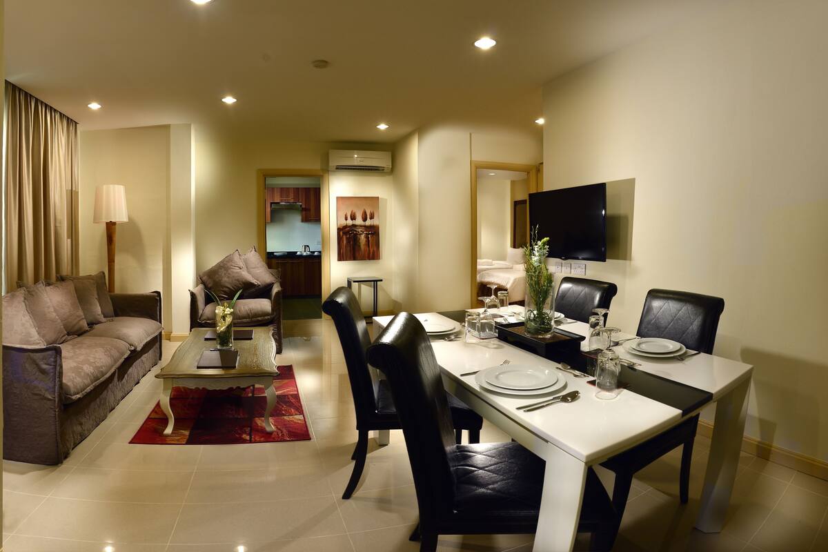 A modern living room with a sofa, a dining table set for four, and a TV mounted on the wall.