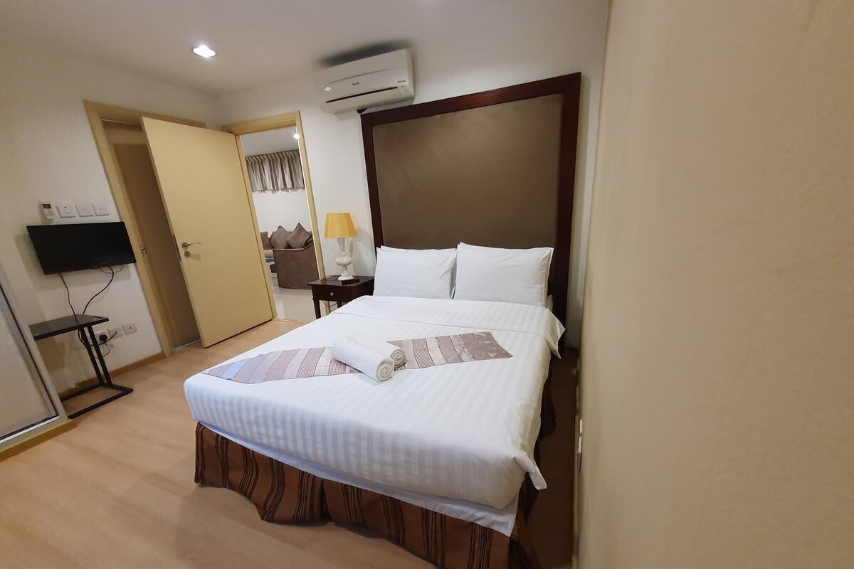 A hotel room with a bed, a TV mounted on the wall, a bedside table with a lamp, and an air conditioning unit on the ceiling.