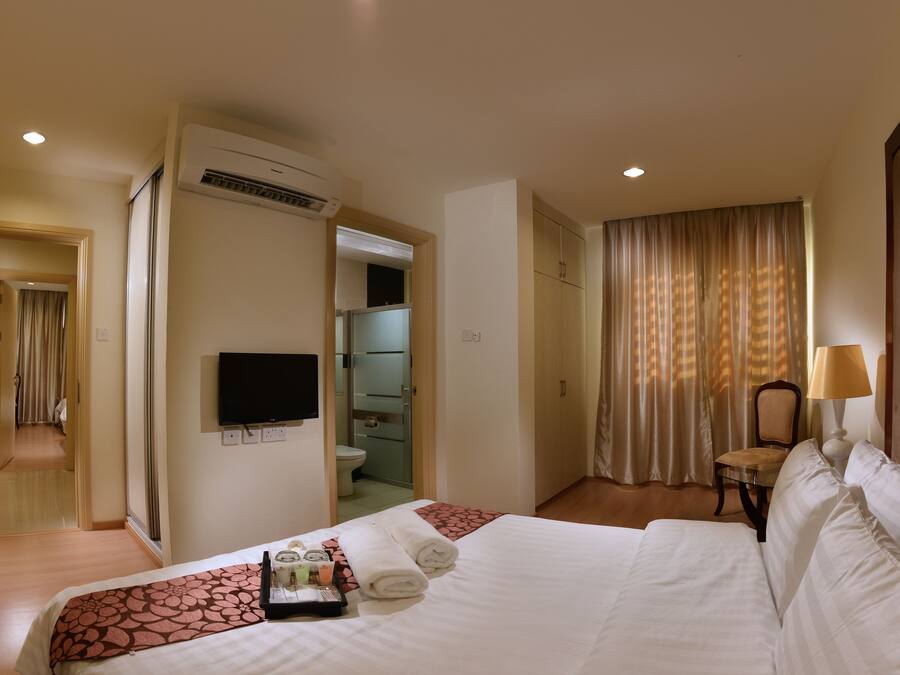 A hotel room with a bed, a TV mounted on the wall, a bathroom visible through an open door, and a chair with a lamp.