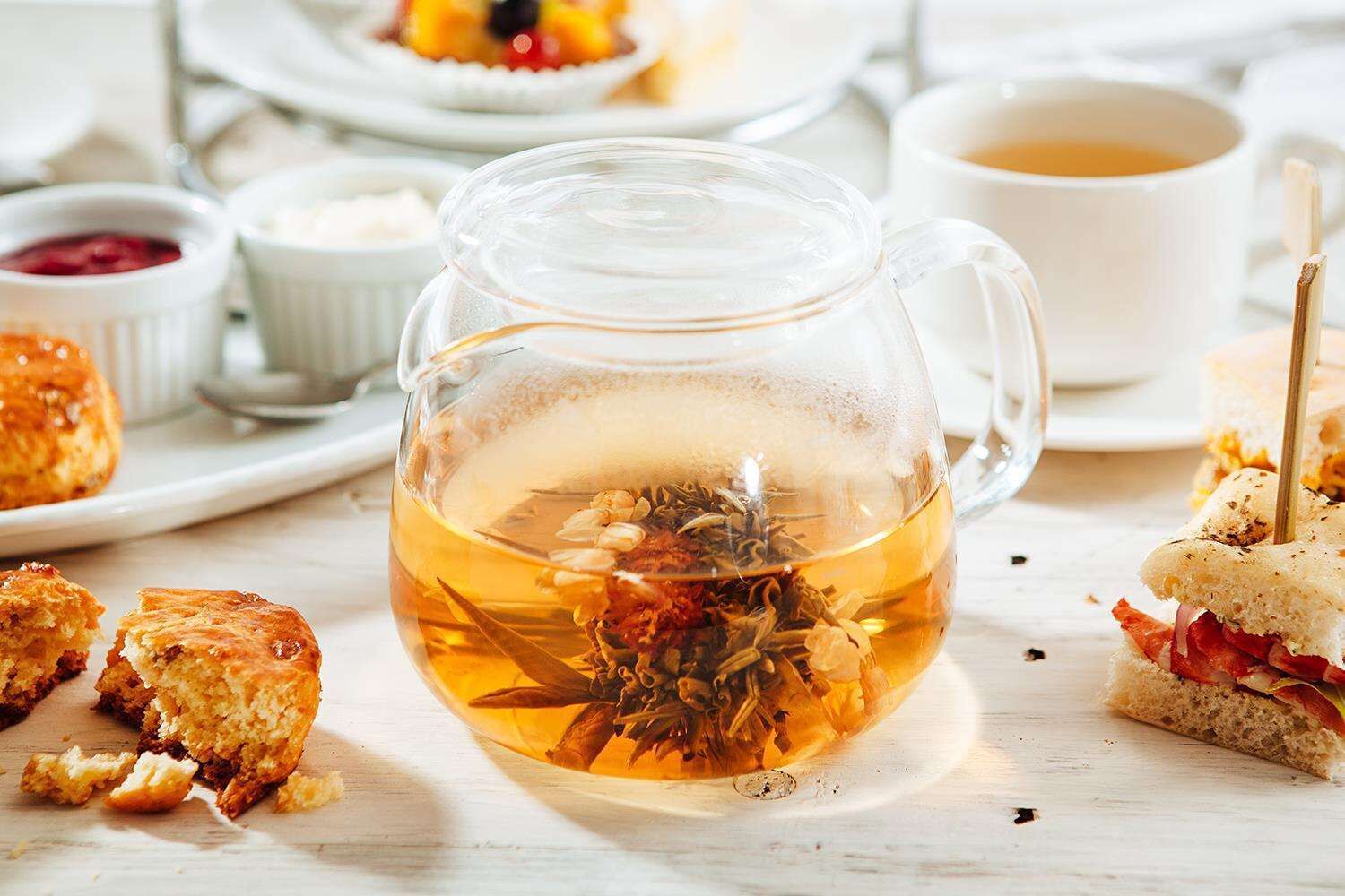 Una taza de vidrio transparente llena de té y hierbas, junto con sándwiches y una taza de té sobre una mesa.