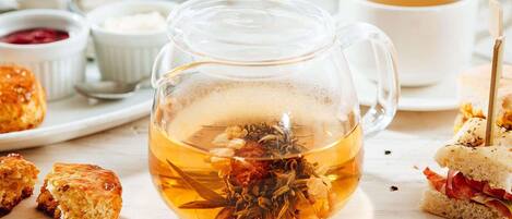 Una taza de vidrio transparente llena de té y hierbas, junto con sándwiches y una taza de té sobre una mesa.