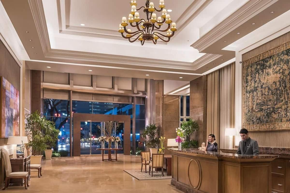 Un vestíbulo de hotel con mostrador de recepción, zona de estar, una gran lámpara de araña y vistas del paisaje urbano a través de puertas de vidrio.