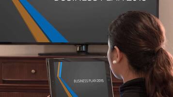 A person holding a tablet displaying 'BUSINESS PLAN 2015' with a TV in the background.