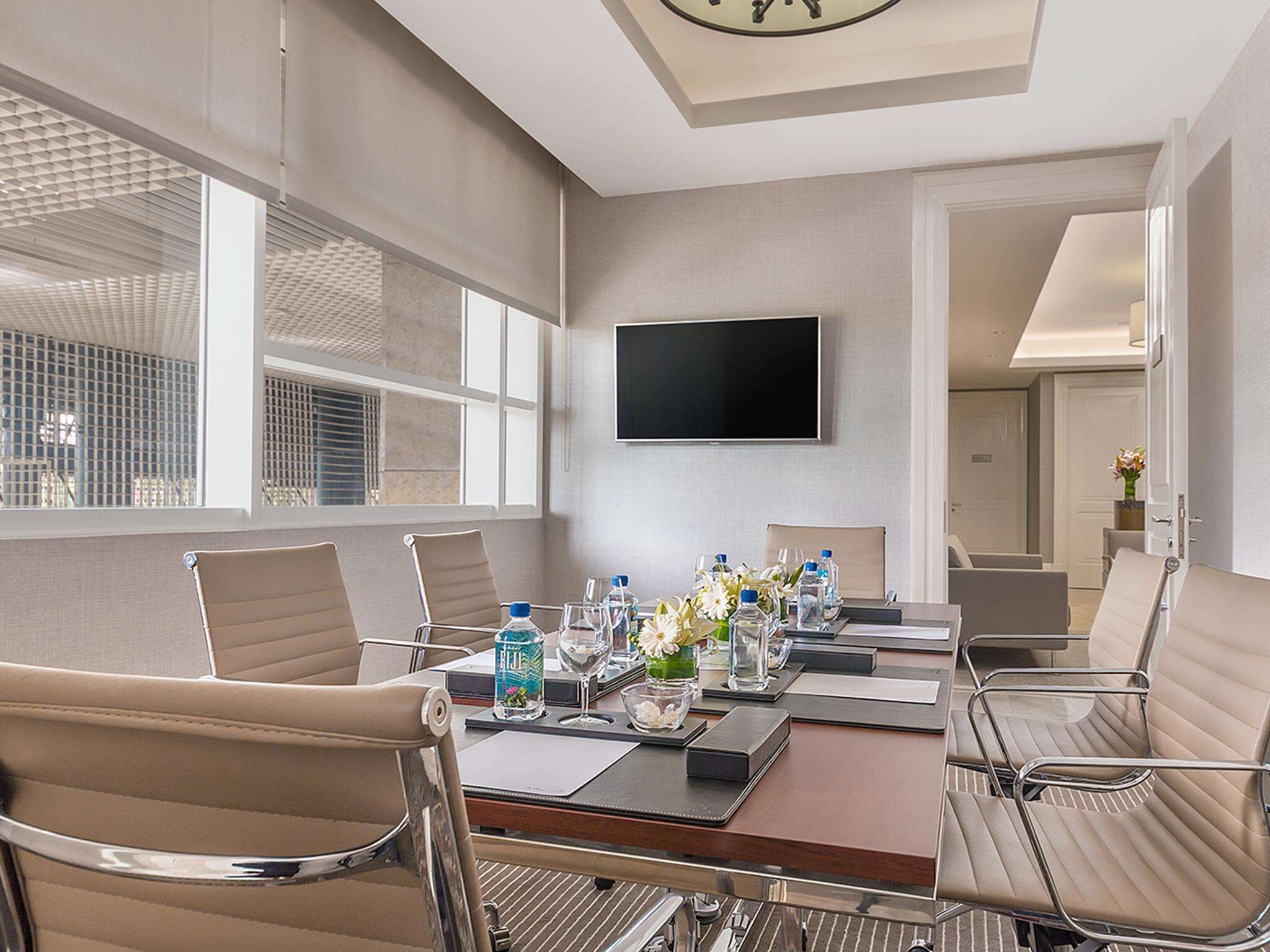 A conference room with a long table, chairs, a flat-screen TV, and a window with blinds.