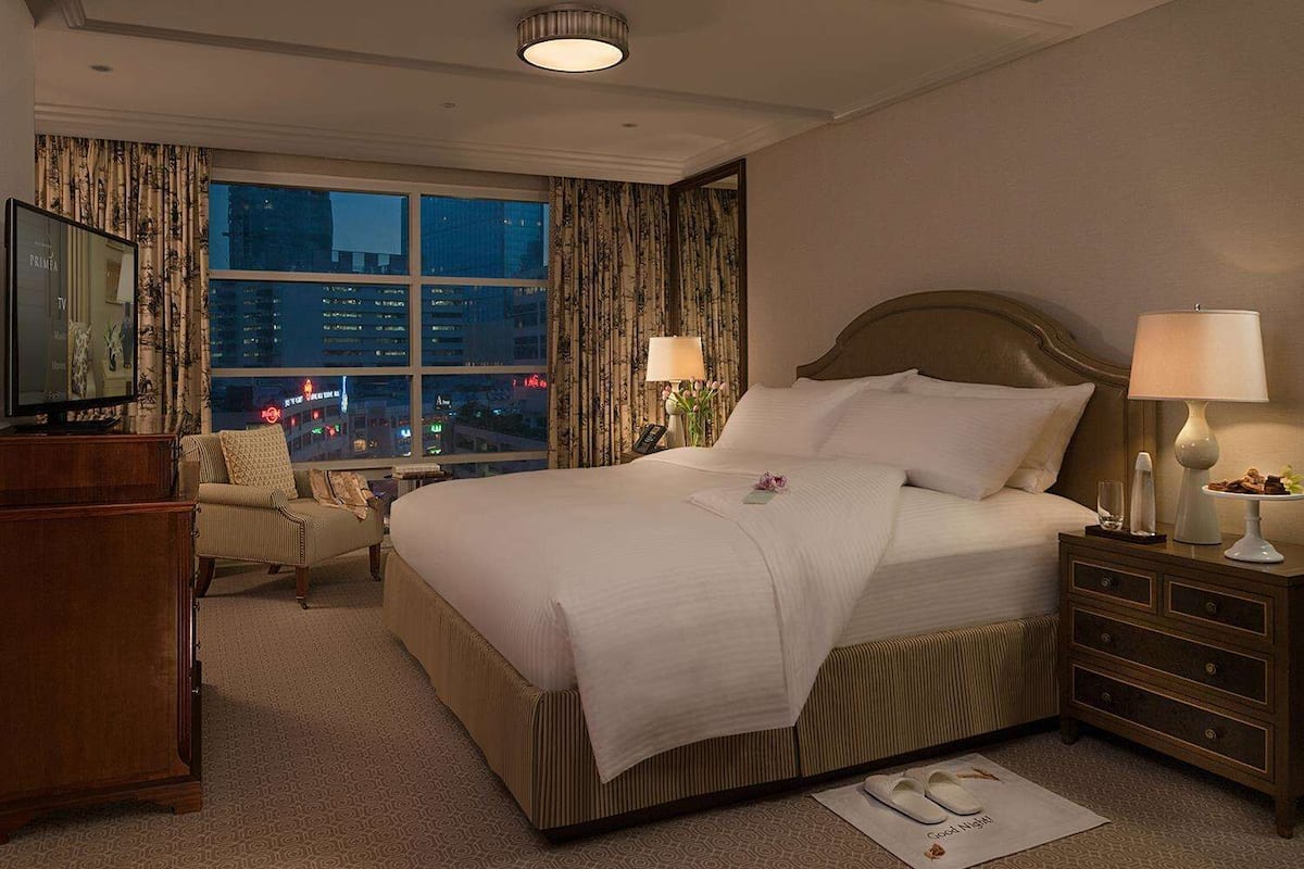Habitación de hotel con una cama grande, televisor, sillón, mesita de noche con lámpara y vista nocturna de la ciudad.