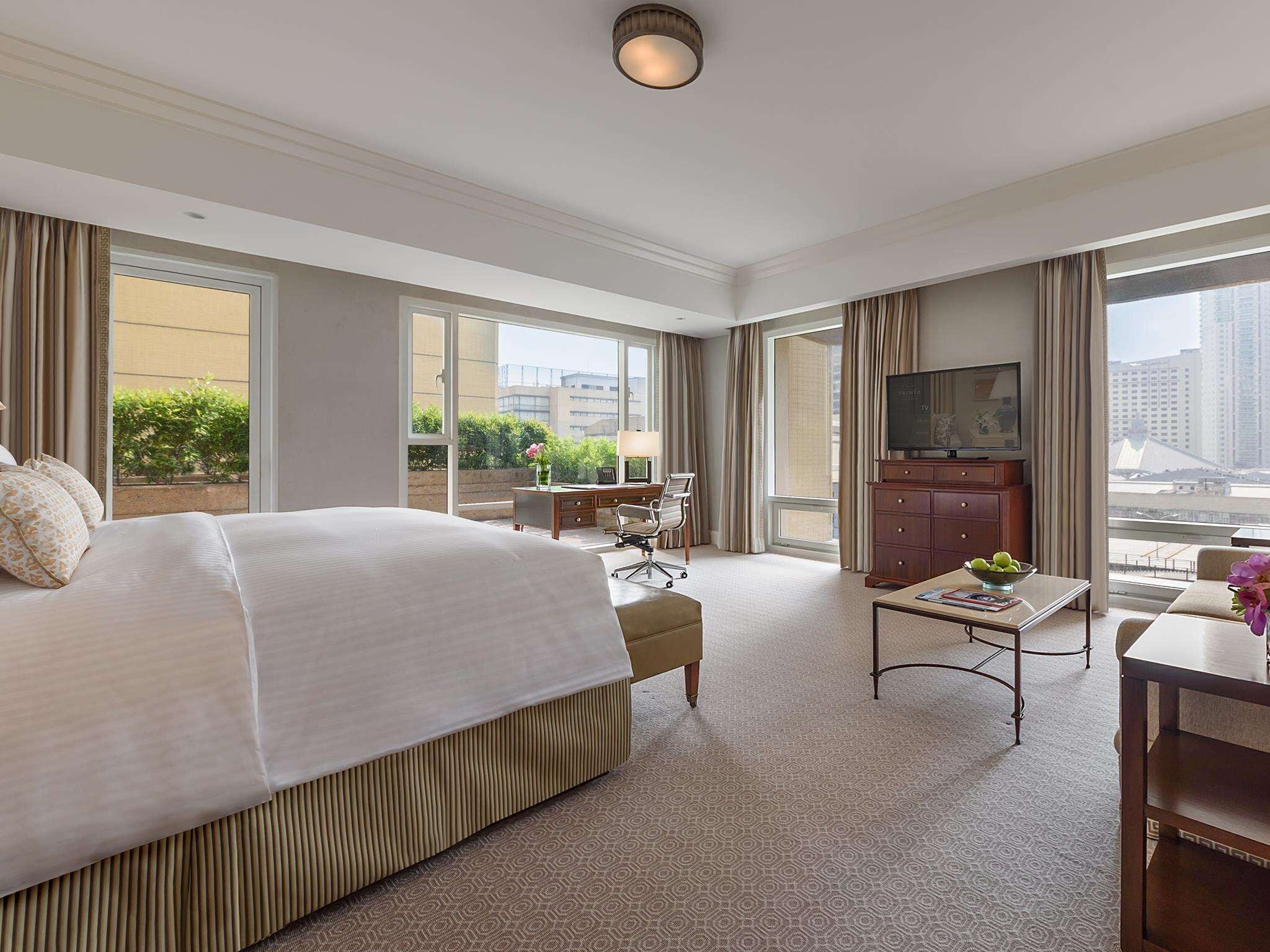 A hotel room with a large bed, a desk with a chair, a television, and a view of the city through large windows.