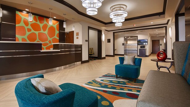 Eine Hotelhalle mit einer Rezeption, Smaragdgrün-Armstühlen, einem grauen Sofa und einem farbenfrohen Teppich.
