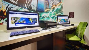 Zwei HP Desktop-Computer auf einem Schreibtisch mit einem grünen Stuhl, einem Drucker und einem Schild für das Business Center an der Wand.