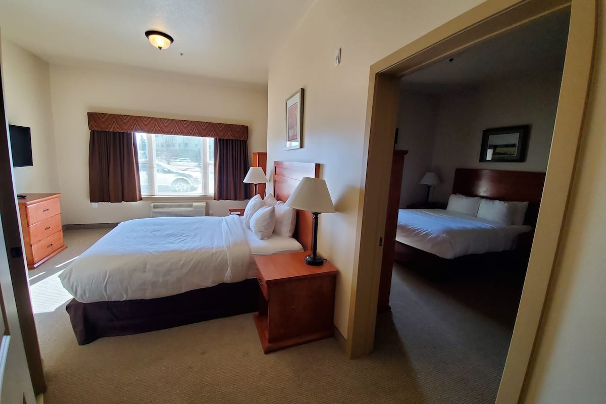 Habitación de hotel con una cama grande, una cama más pequeña, una mesita de noche con lámpara, un aparador y una ventana con cortinas.