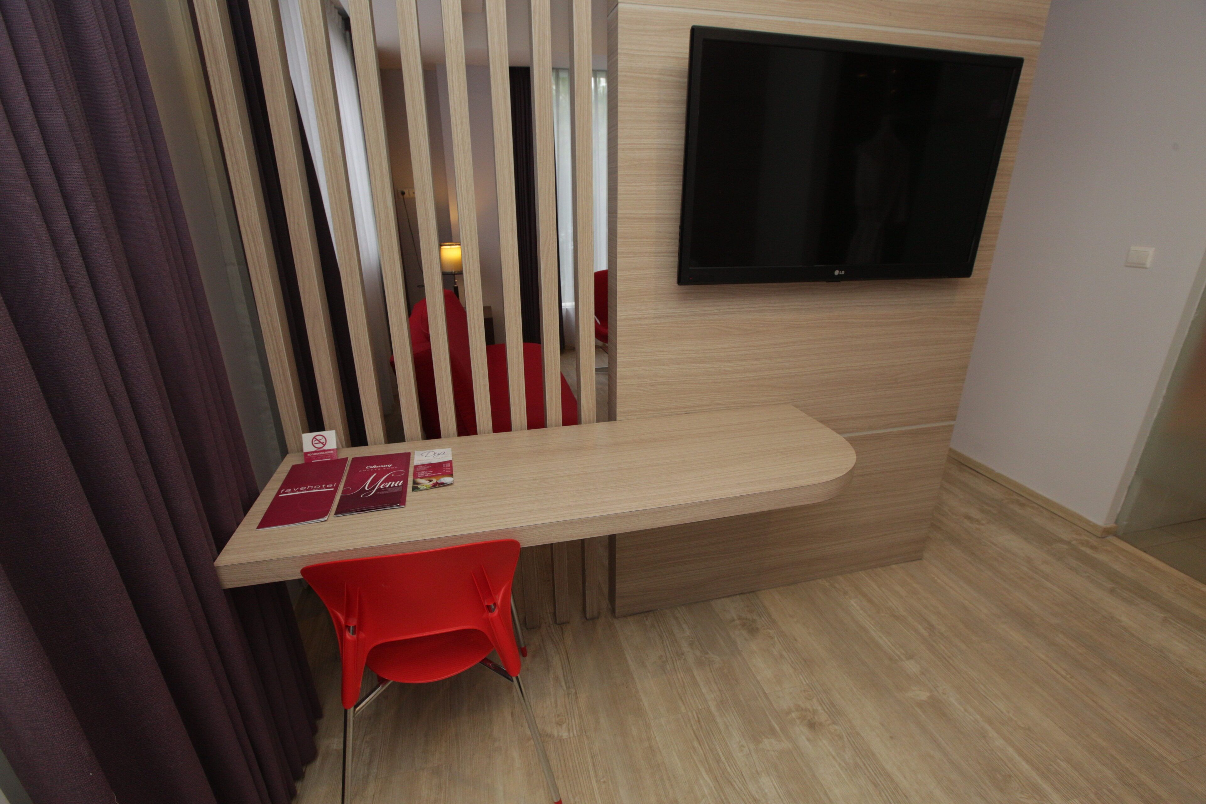 A modern room with a wooden desk, a red chair, a flat-screen TV mounted on the wall, and a window with curtains.