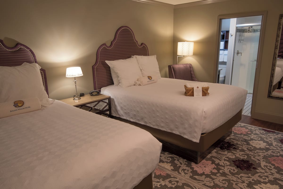A hotel room with two beds, a nightstand, a lamp, and a bathroom visible through an open door.