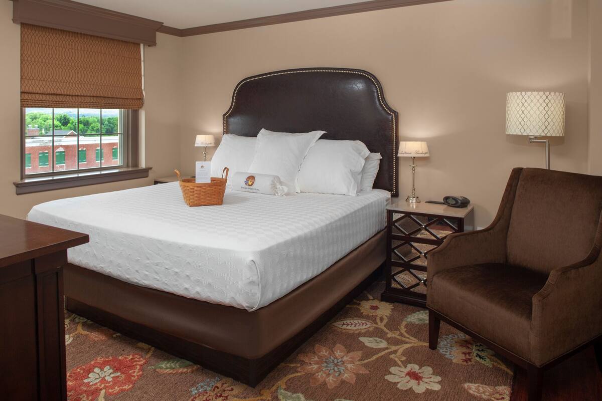 A hotel room with a large bed, a brown armchair, a nightstand with a lamp, and a window with a view of buildings.